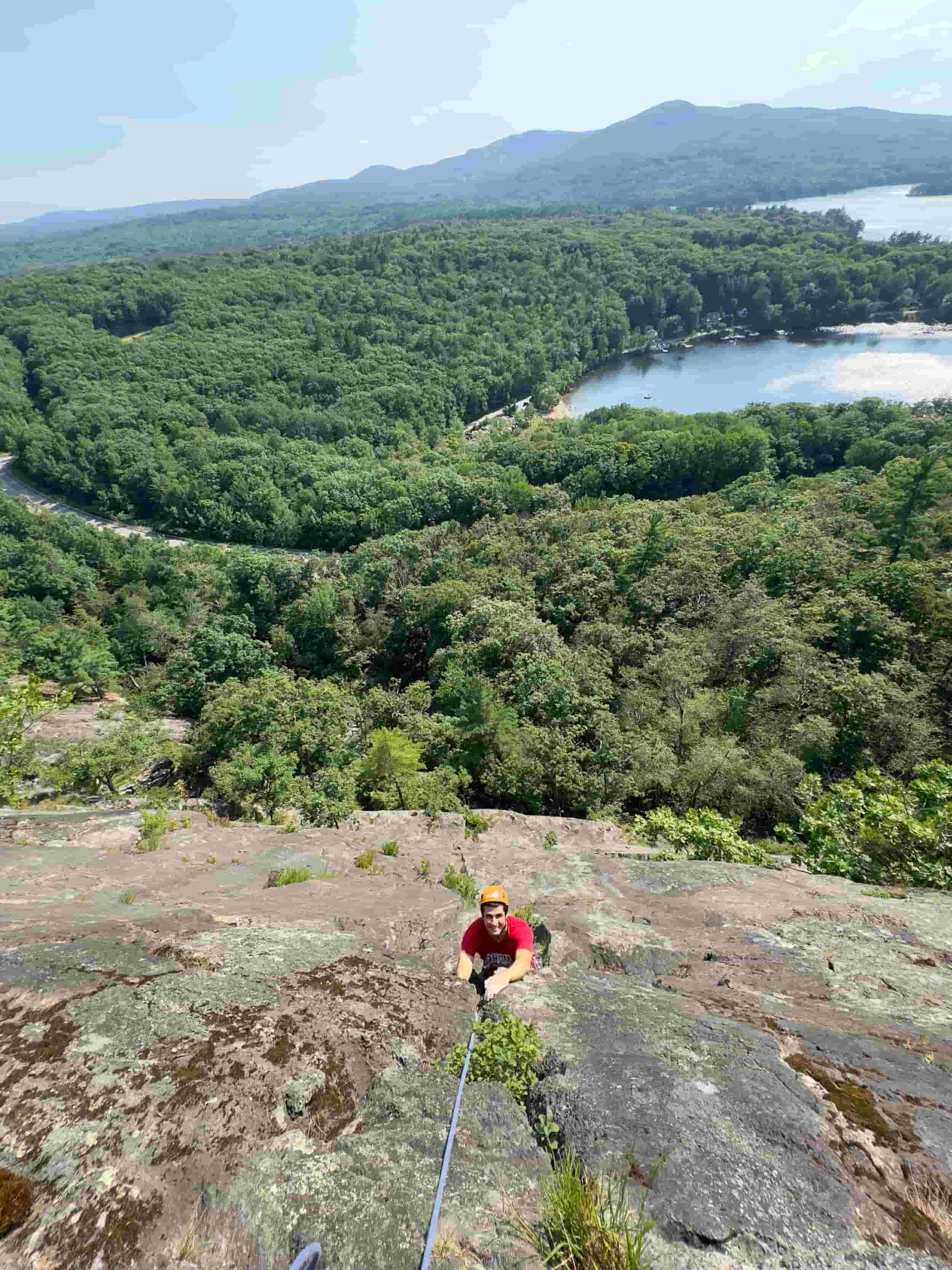 A person rock climbing on a steep cliffside with a forested landscape, a lake, and mountains in the distance.