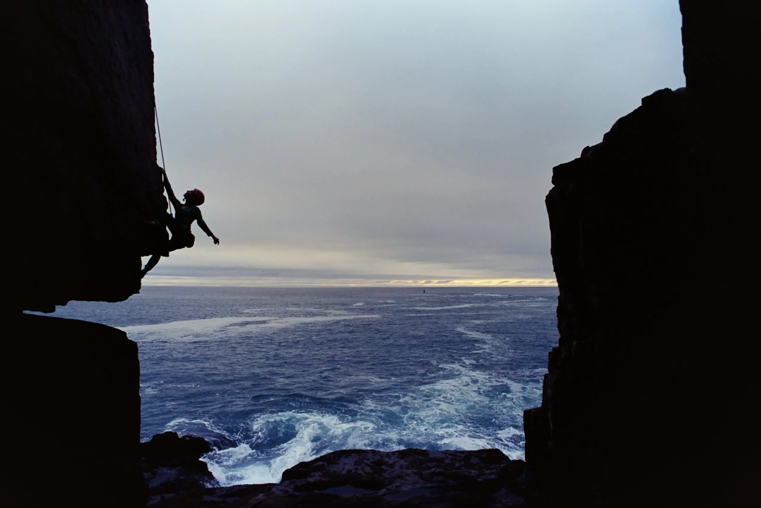 A person rock climbing on a cliff with an ocean view in the background, seen through a natural rock opening during dusk or dawn.