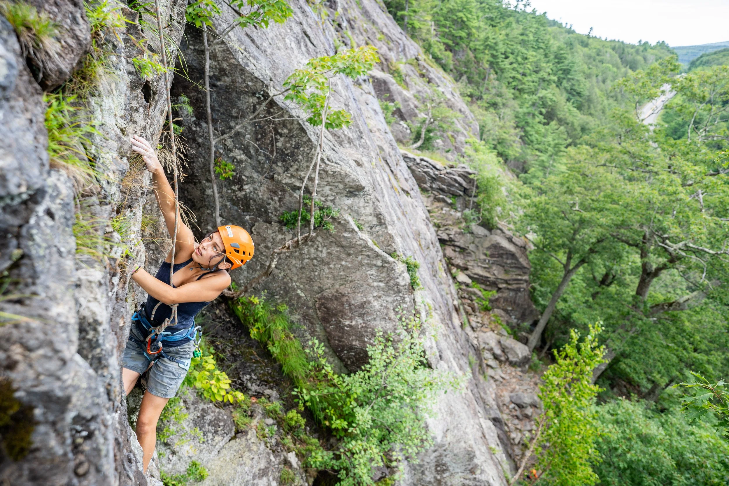 A woman rock climbing outdoors on a steep, rocky cliff with green trees in the background, wearing a helmet and climbing gear.