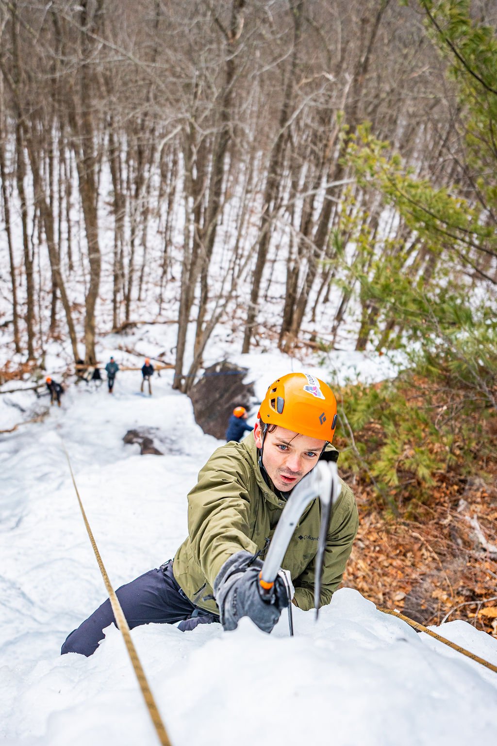 A person in a green jacket and orange helmet climbing a snowy ice wall with crampons and ice axes, using safety ropes, in a forested area.
