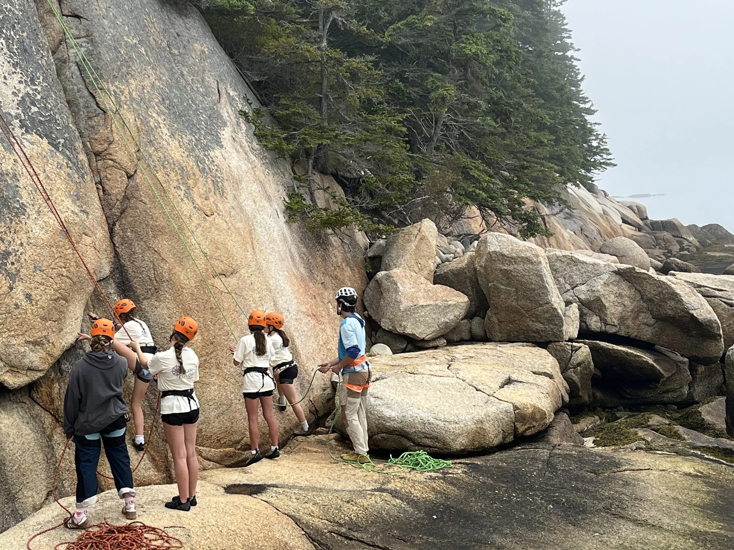 Group of people wearing helmets and harnesses preparing to climb a rock face on a foggy day at the beach.