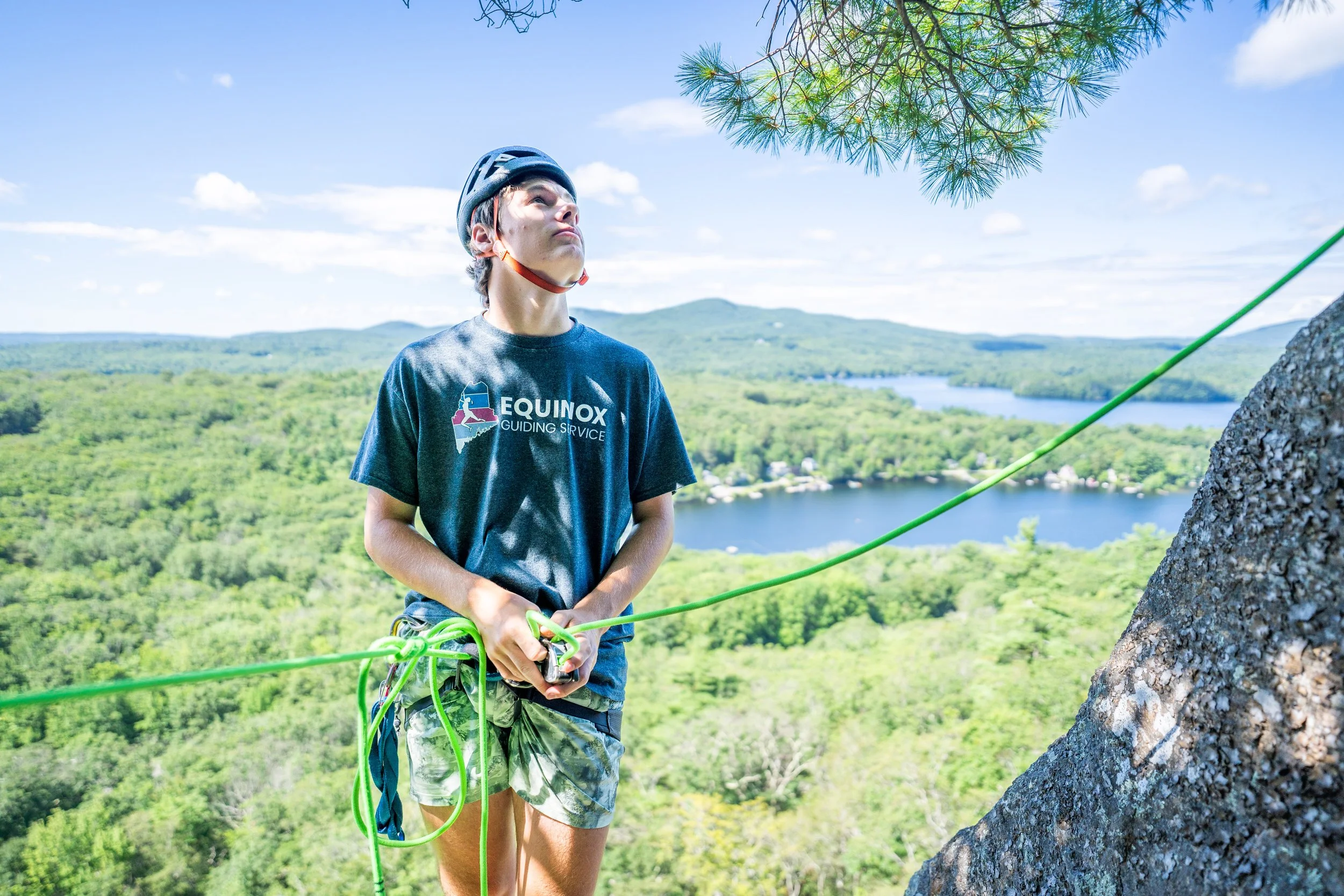 A person wearing a helmet and a T-shirt with 'EQUINOX GUIDING SERVICE' is rock climbing on a high outdoor cliff with a view of a lush green forest and lakes below, under a partly cloudy sky.