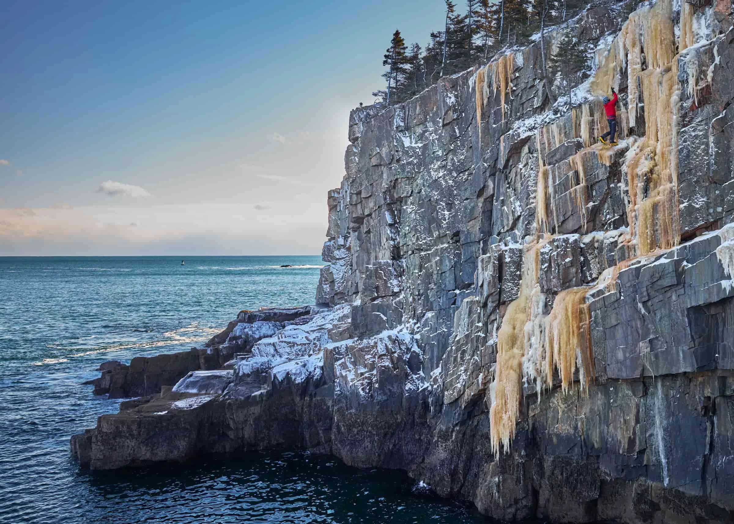 Person ice climbing on a snowy, frozen waterfall on a rocky cliff beside the ocean.