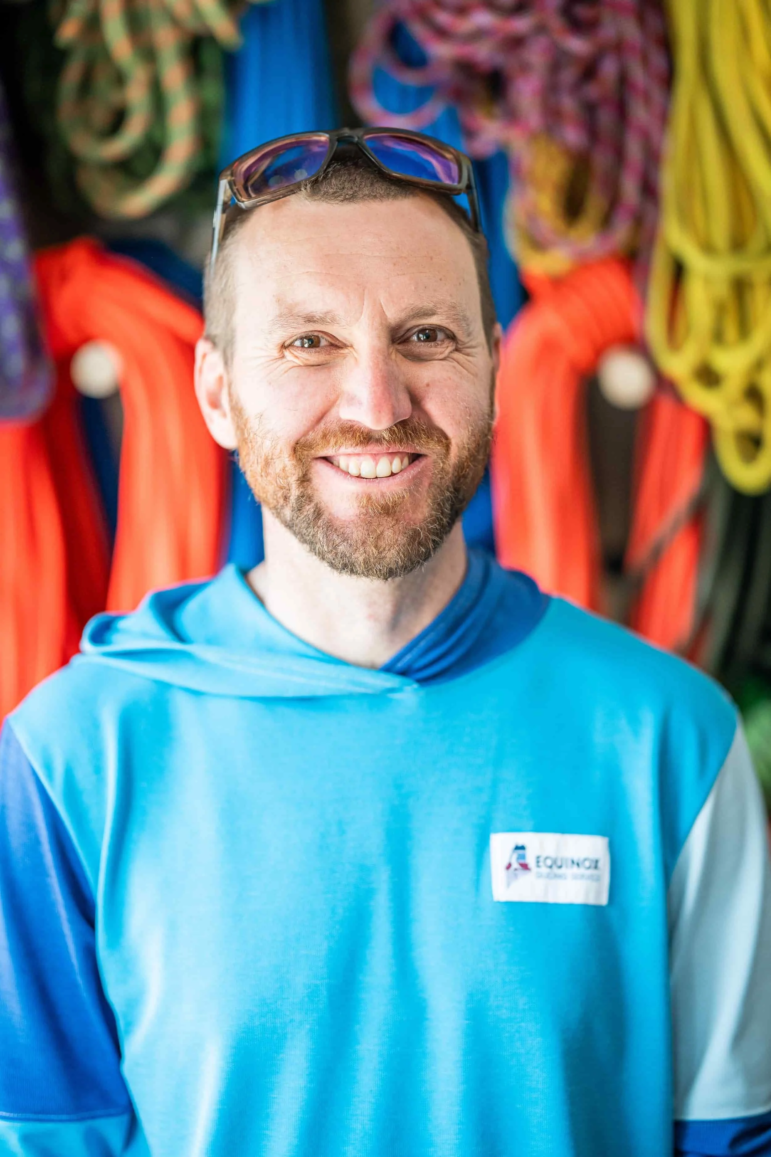 A smiling man with a beard and mustache, wearing sunglasses on his head and a blue shirt, standing in front of climbing ropes and gear.