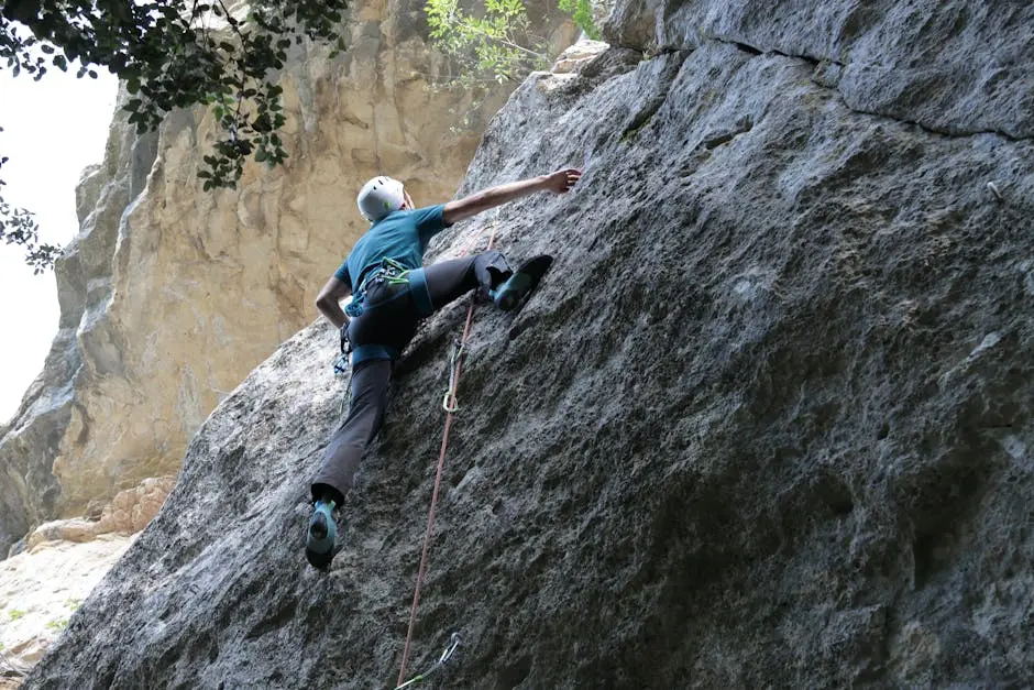 A person rock climbing on a steep outdoor cliff, wearing a helmet, harness, and climbing shoes, with safety rope attached.