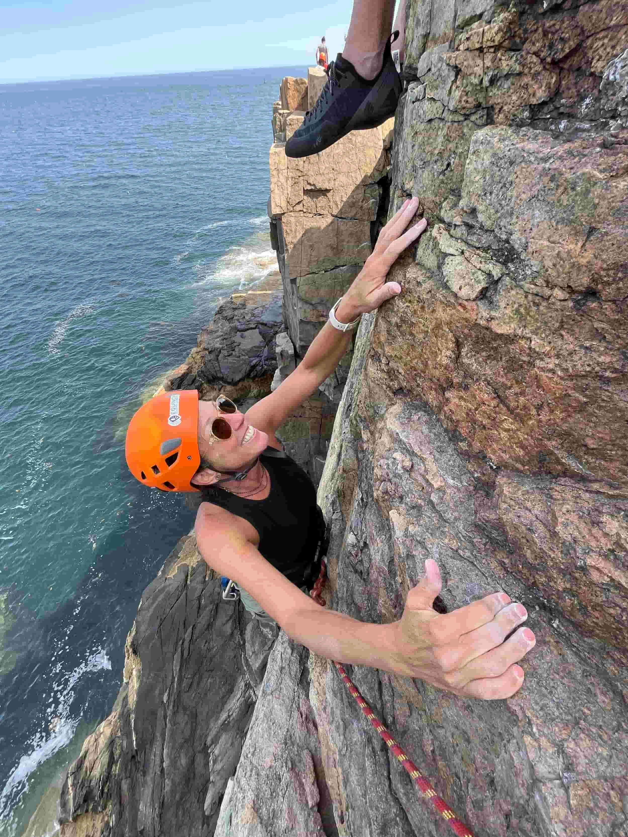 Woman rock climbing on a cliff near the ocean, wearing an orange helmet and sunglasses, smiling and giving a thumbs-up.