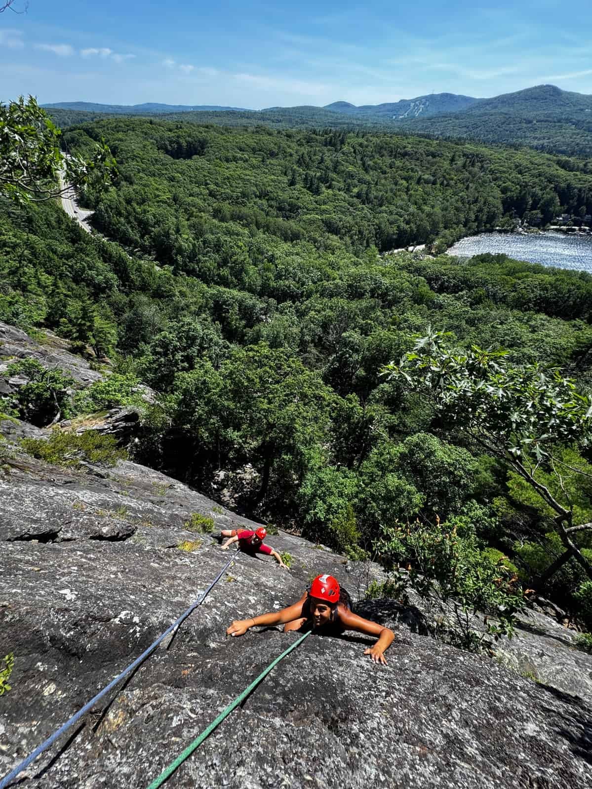 Two people wearing helmets and climbing gear rock climbing on a steep outdoor rock face with a lush green forest and a lake in the background.
