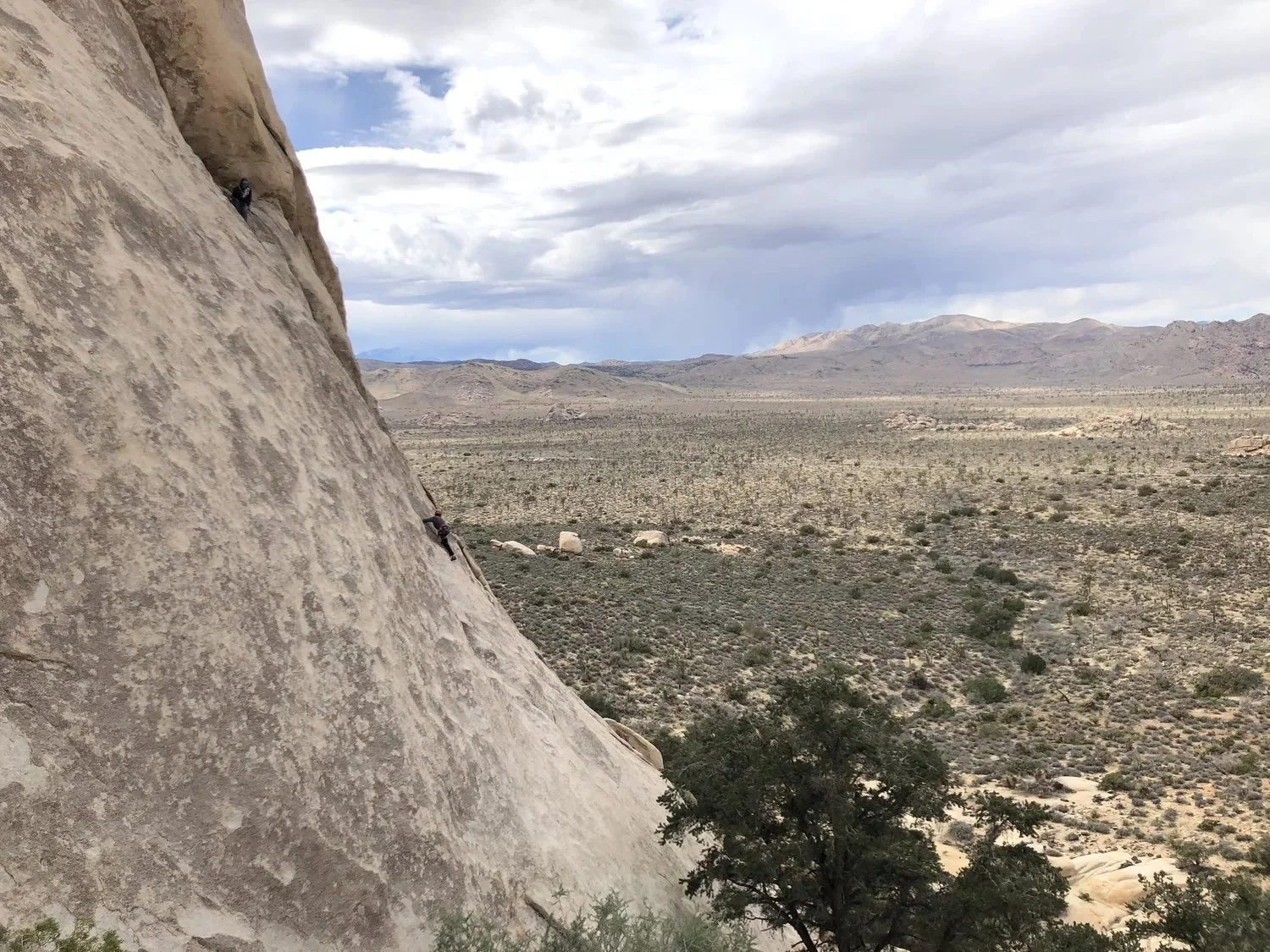 Two climbers ascending a steep, large rock face in a desert landscape with mountains and cloudy sky in the background.