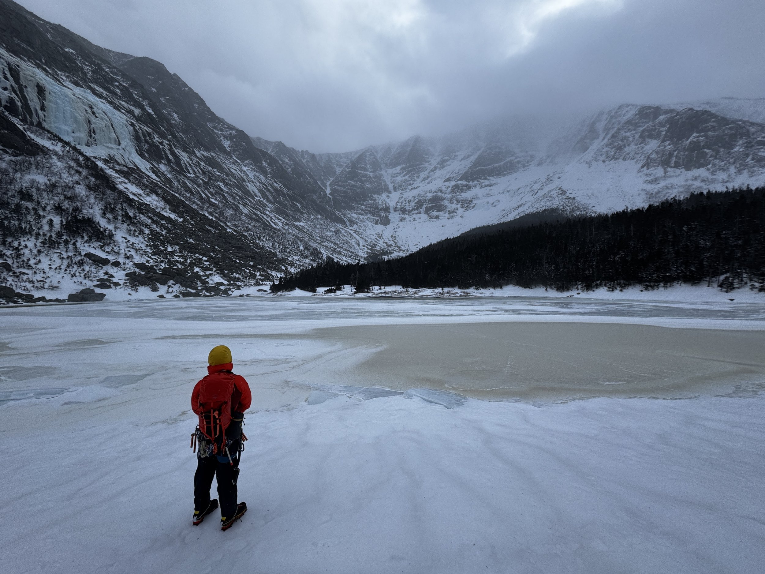 A person dressed in red and yellow winter gear stands on snow-covered ground facing a frozen lake surrounded by snow-capped mountains and pine trees.