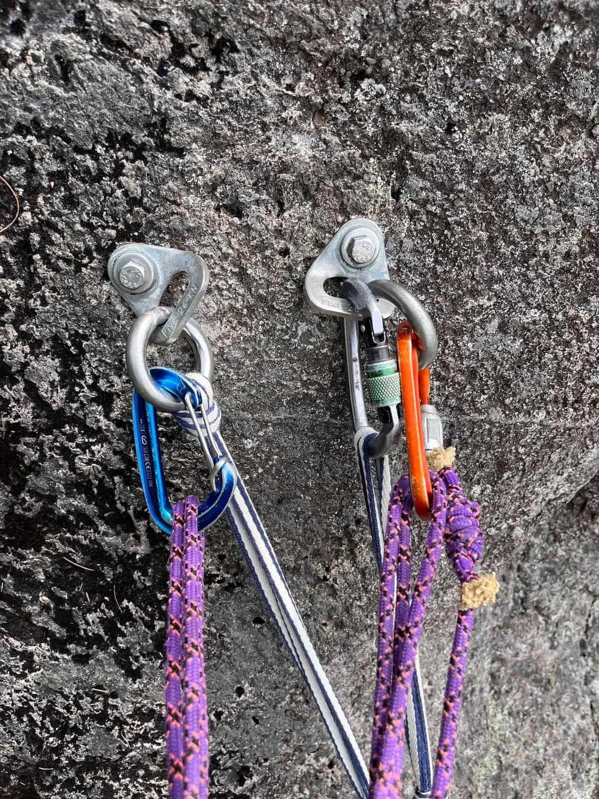 Close-up of two climbing anchors attached to a rock wall. Each anchor has carabiners, one blue and one orange, connected to purple climbing ropes.