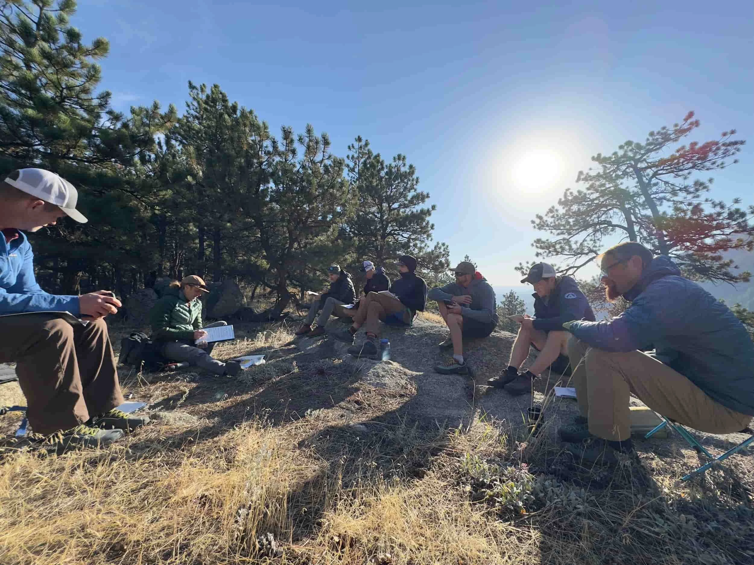 A group of people sitting on rocks and logs outdoors in a forested area, with some reading and others using electronic devices, under a clear blue sky with the sun shining.