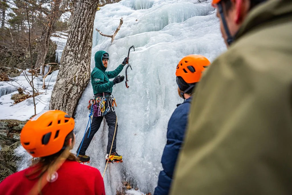 Ice climber with equipment demonstrating ice axes to group during ice climbing session on a frozen waterfall, in winter outdoor setting.