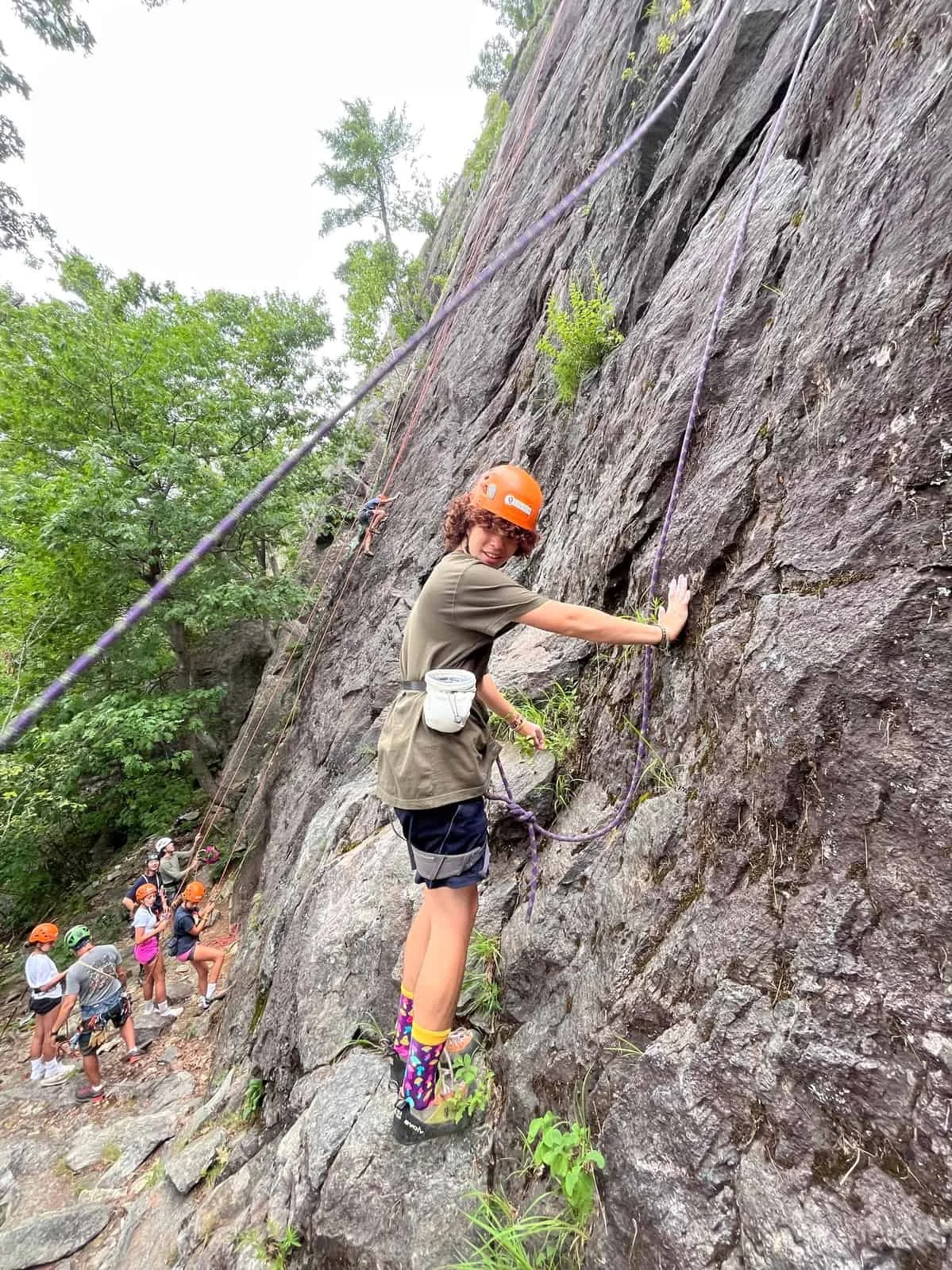 A group of children rock climbing outdoors, with one girl in the foreground wearing a harness, helmet, colorful socks, and smiling while holding onto the rock face, and others waiting in line behind her.
