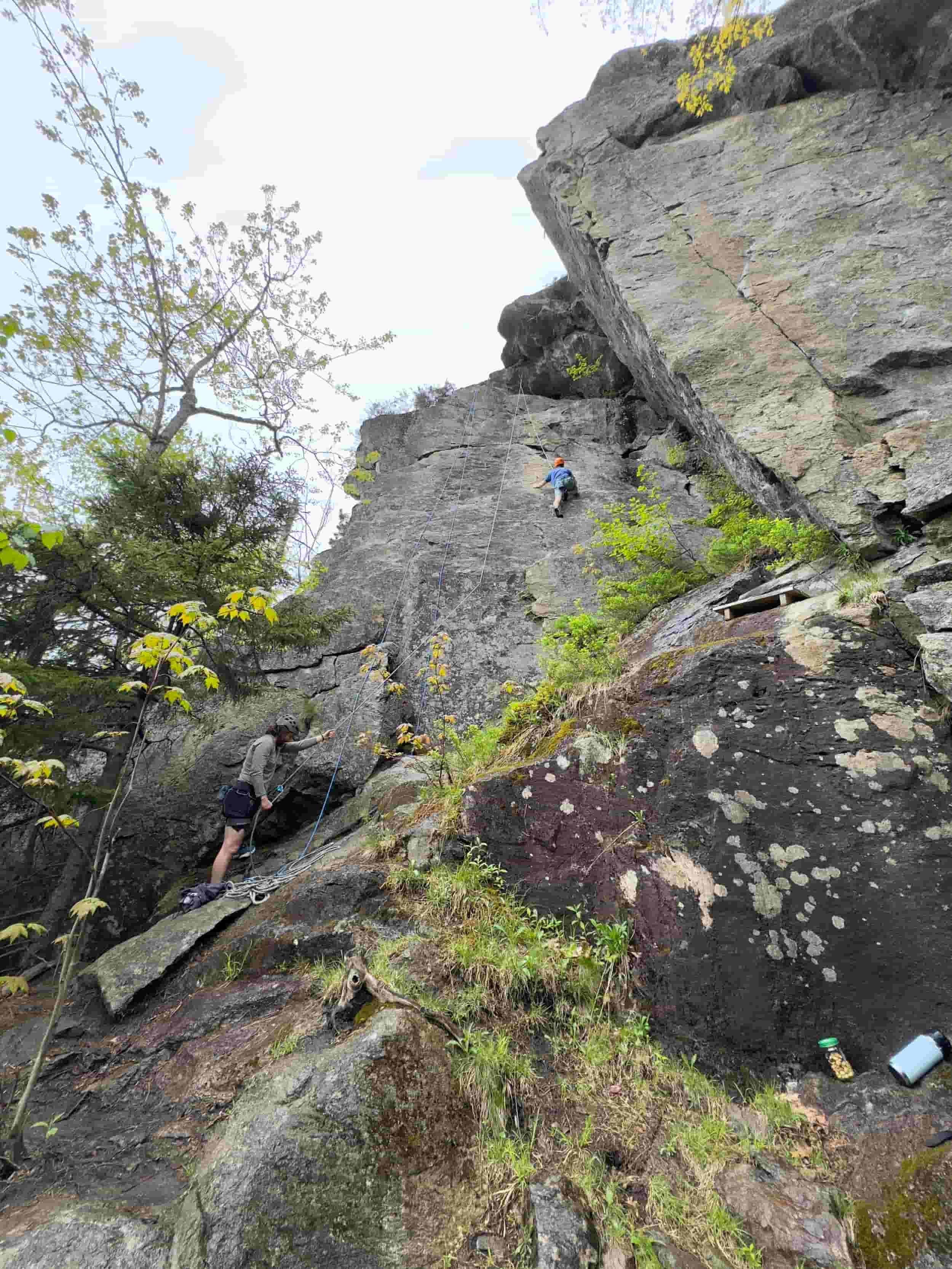 Two climbers are ascending a steep rock face with the help of ropes, one person near the bottom and the other higher up, surrounded by trees and rugged terrain.