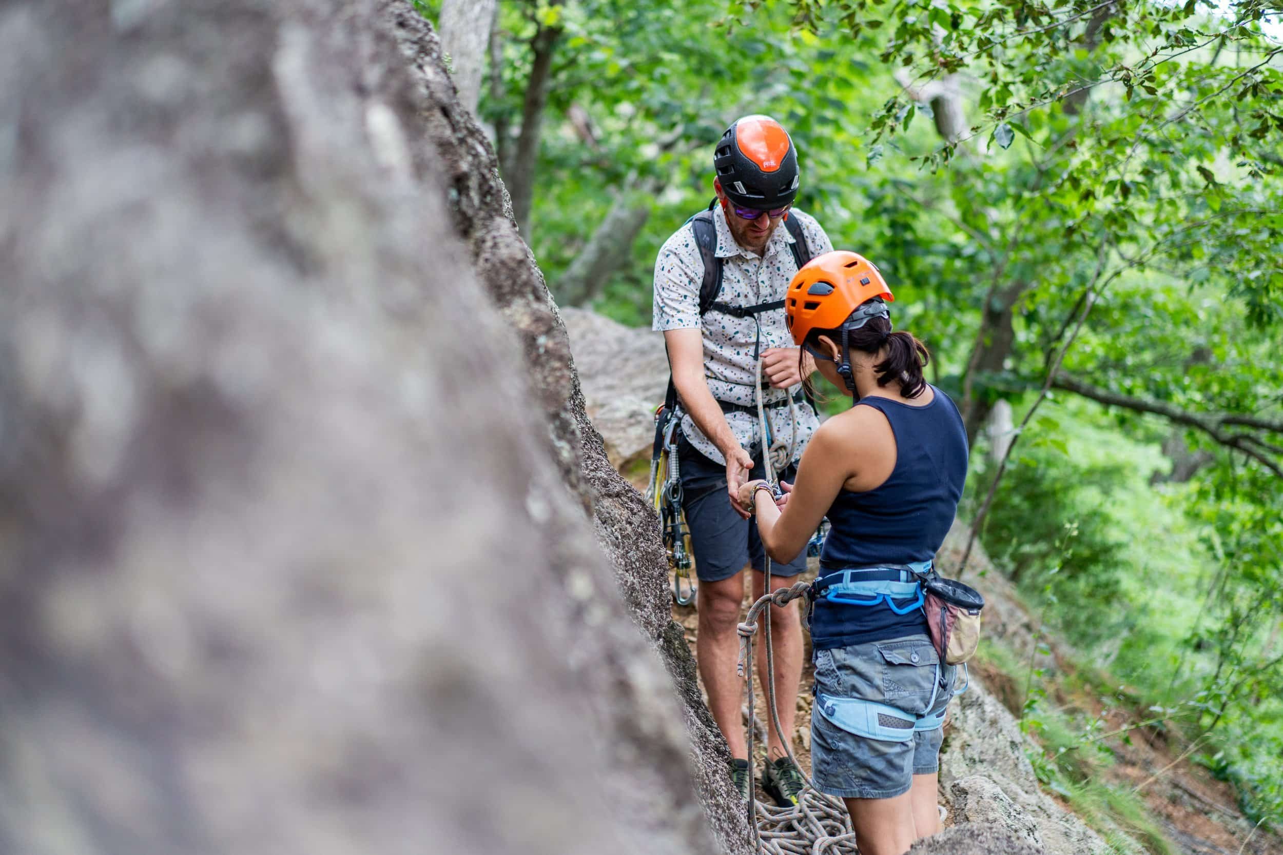 Two climbers in helmets on a rocky outdoor trail securing climbing gear together, surrounded by green trees.
