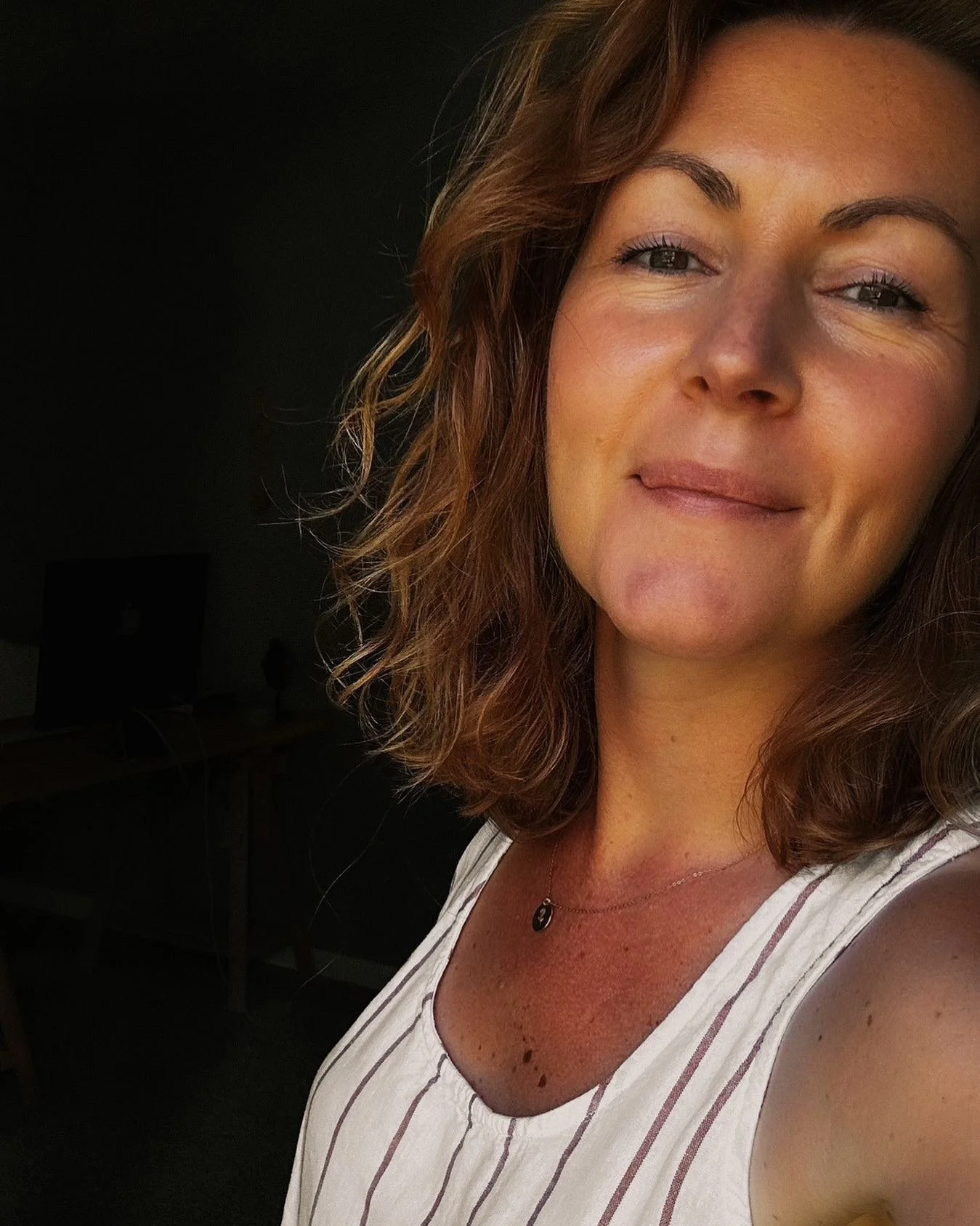 A woman with shoulder-length wavy brown hair, light skin, and freckles wearing a sleeveless striped top with a small necklace taking a selfie indoors near a window with sunlight on her face.