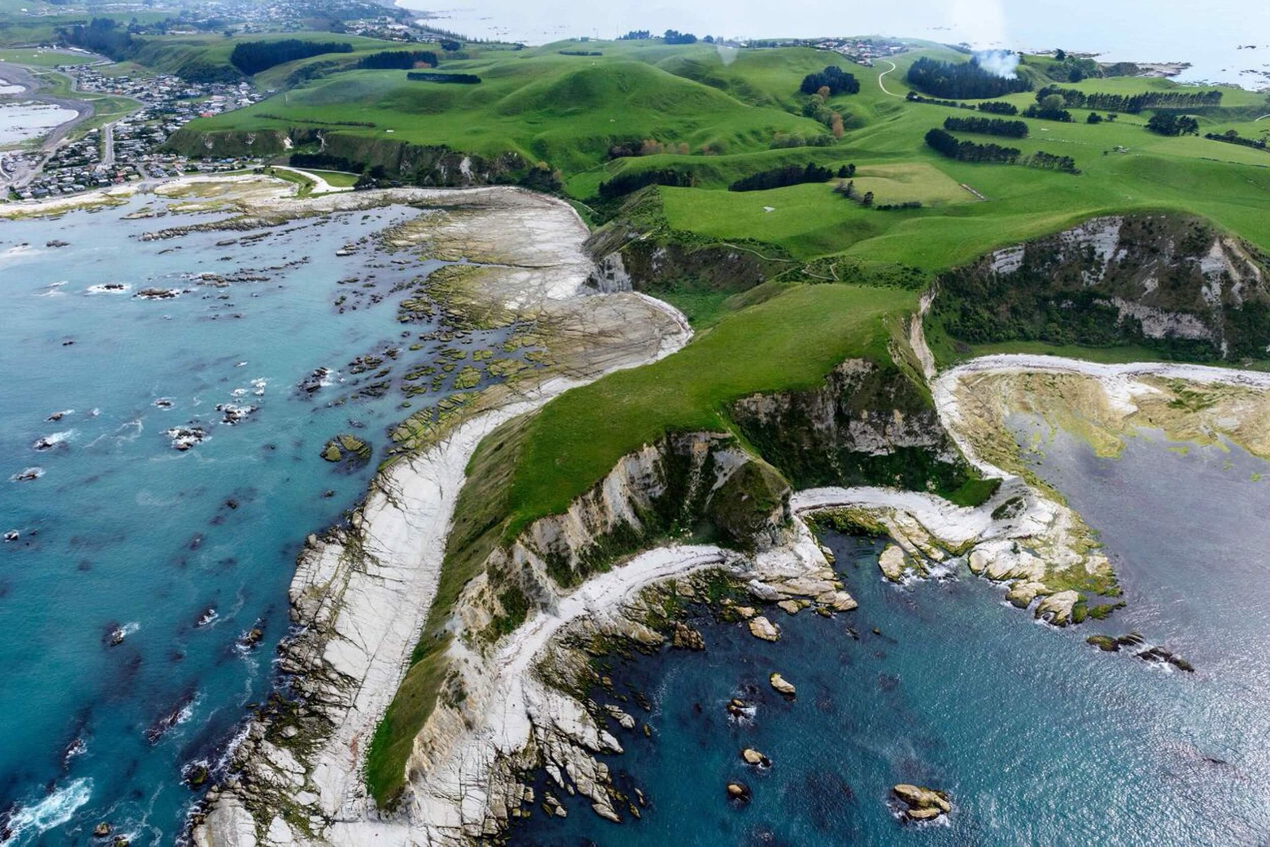 The aerial view of Whalers Bay in New Zealand showcases a stunning coastal landscape with white cliffs and rolling green hills, divided by the deep blue of the sea and dotted with rocky islets.