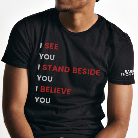 Close-up of a person wearing a black T-shirt with red and white text that says, "I see you, I stand beside you, I believe you."