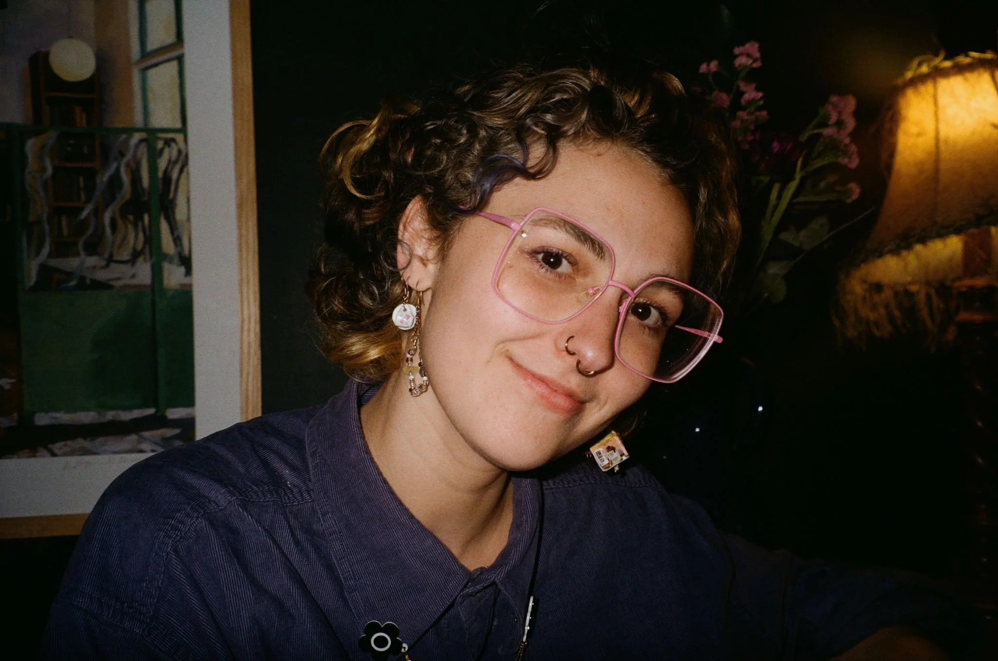 A young woman with curly brown hair and dark eyes smiles at the camera. She is wearing a blue shirt, pink square glasses, two nose rings and earrings. Her head is tilted at an angle.