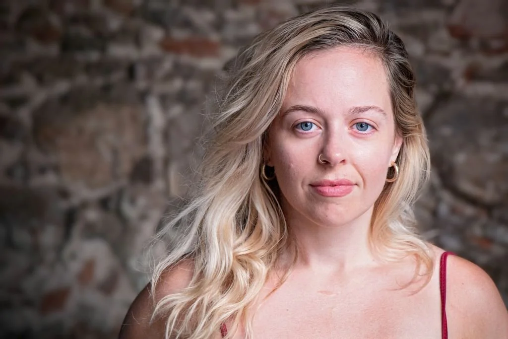 Close-up portrait of a young woman with long blonde hair, blue eyes, wearing gold hoop earrings and a gold nose ring, and a red strap top, standing against a stone wall