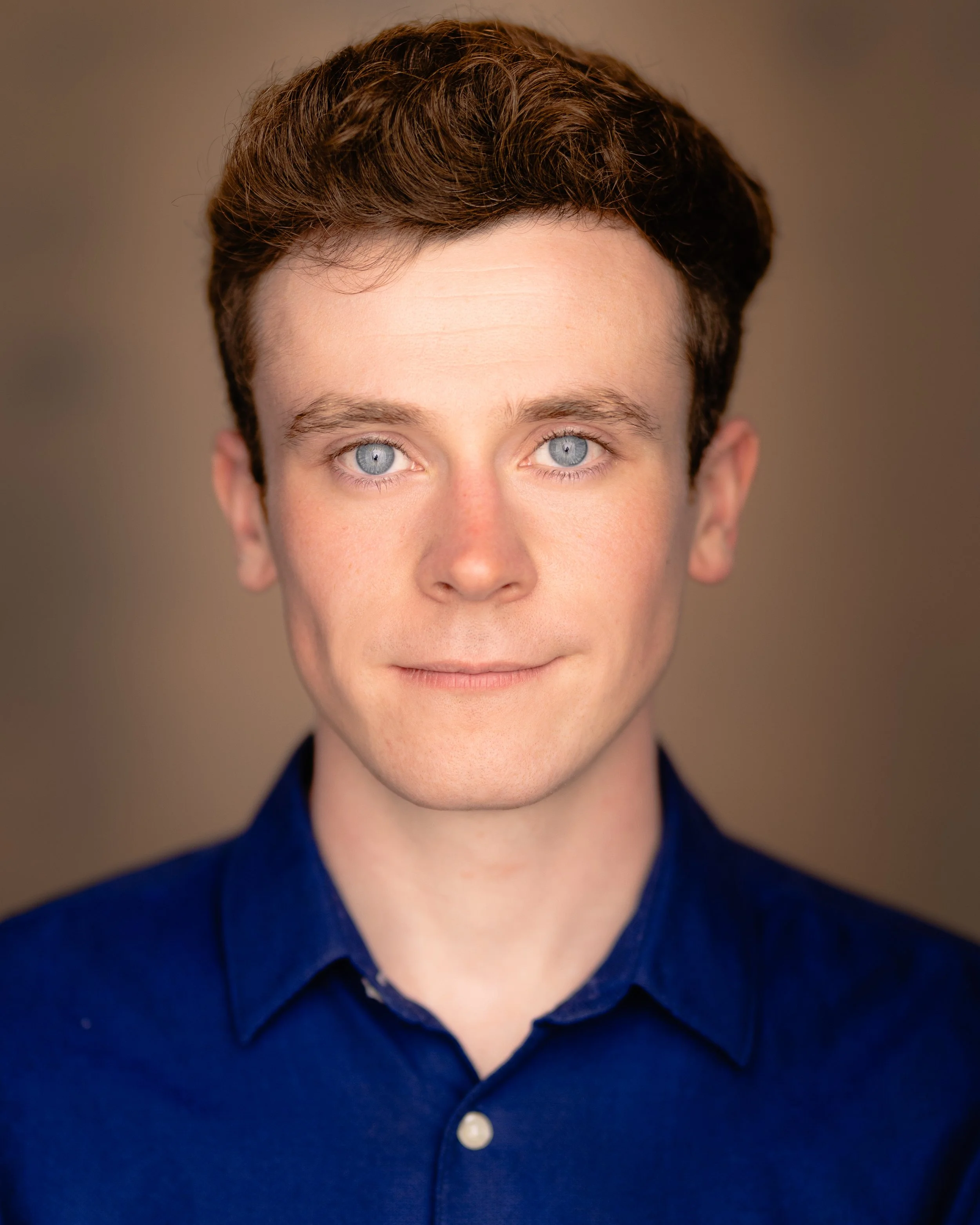 A man with brown hair and blue eyes, wearing a blue shirt, looks directly at the camera. He is clean shaven and has light skin. He is against a brown background