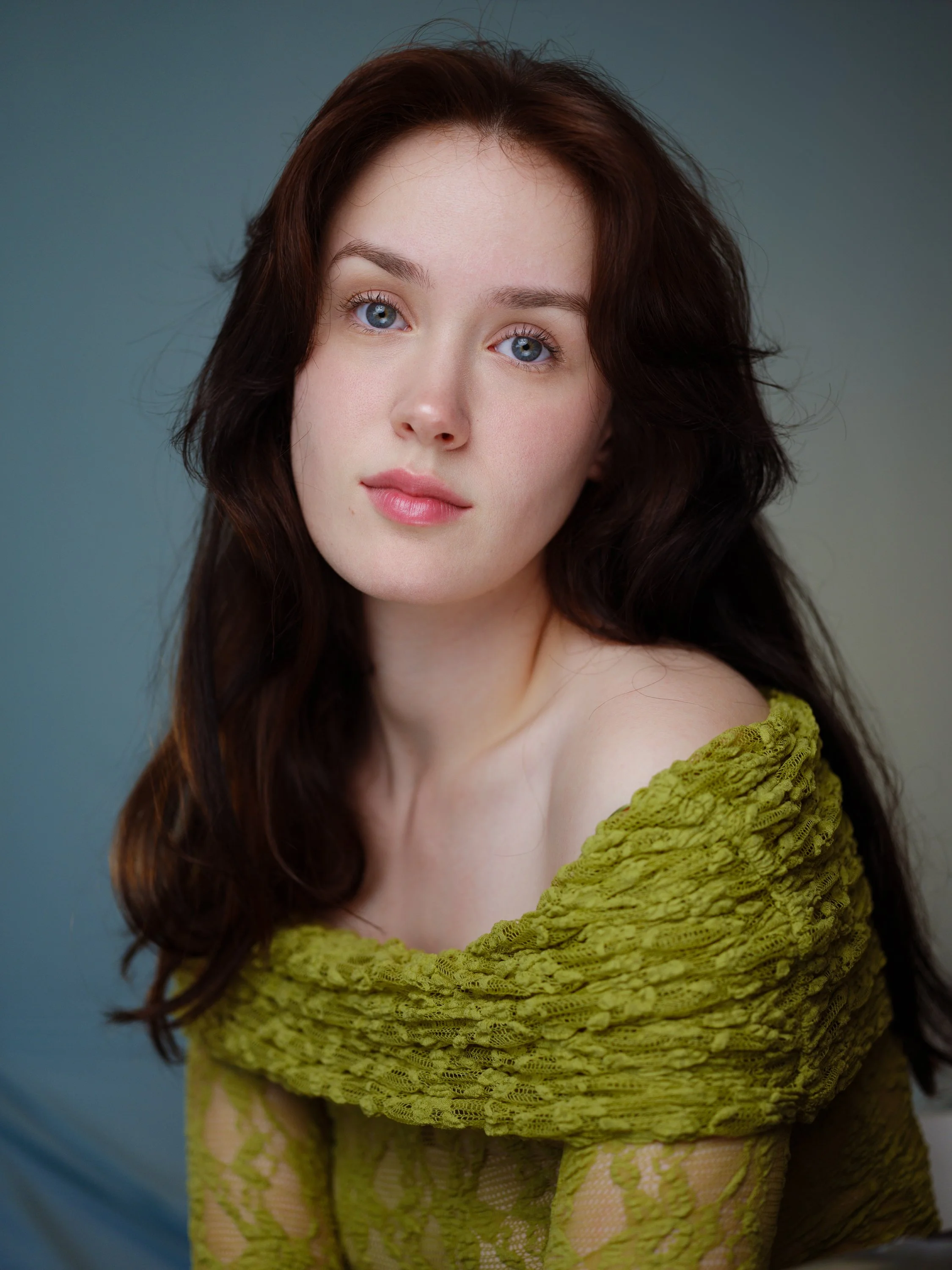 A young woman with long dark hair, pale skin and blue eyes, looks at the camera - she is sitting at an angle. She's wearing an olive green off-the-shoulder lace top and the background is dark grey
