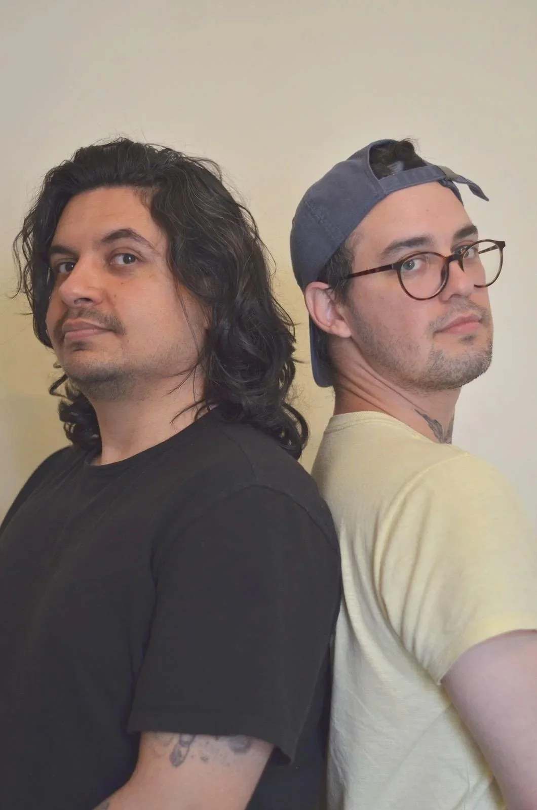 Two young men stand back to back looking at the camera. The man on the left has dark shoulder length hair, dark eyes, brown skin and a moustache, and is wearing a black tshirt. The man on the right has brown hair and light skin, with glasses & a cap