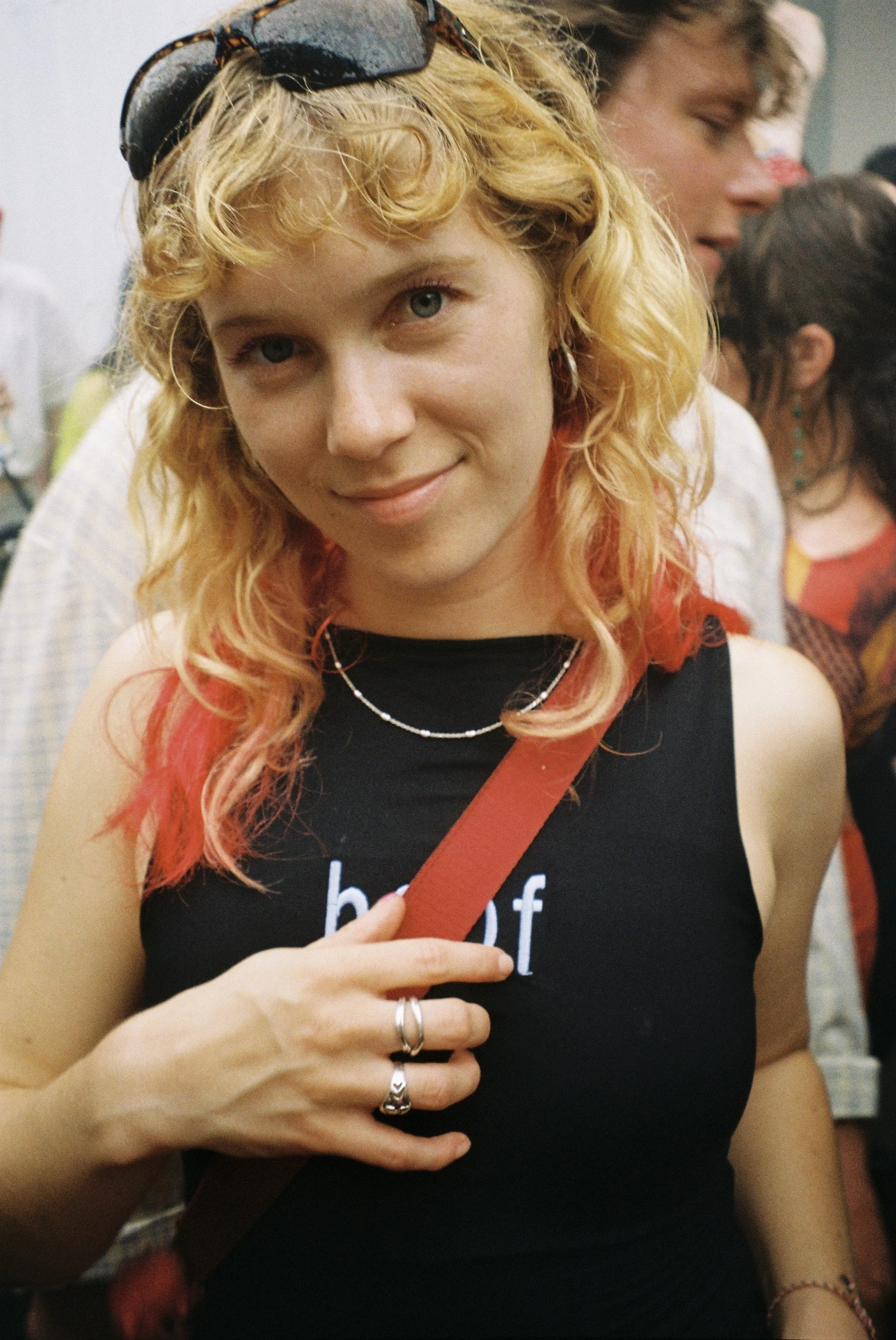 A young woman with light skin, curly blonde hair, a fringe and blue eyes, smiles at directly at the camera. She is wearing a black vest top, a red strap bag, sunglasses on her head, and gold earrings and necklace.