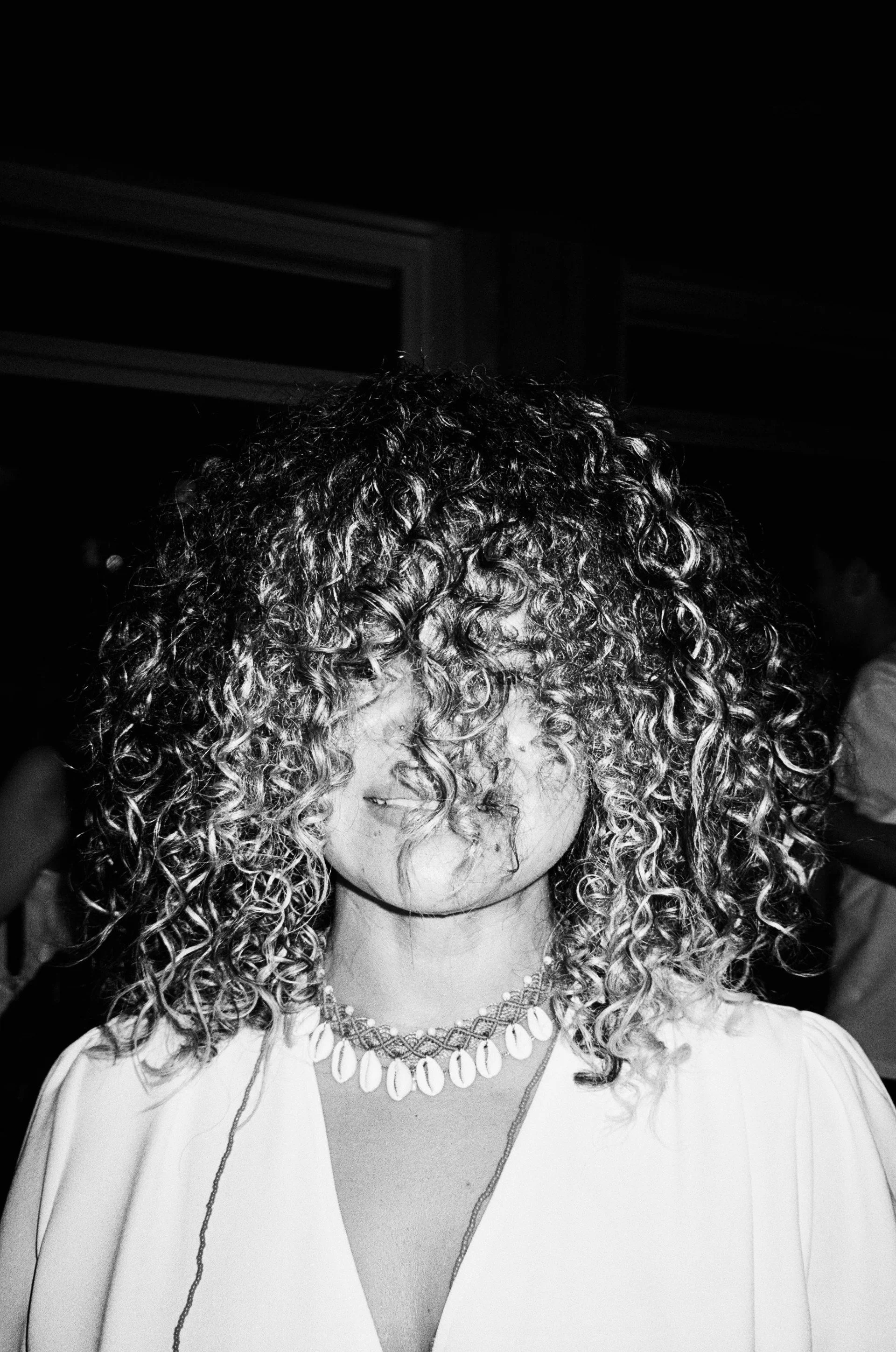 A black and white photo of a young woman with curly hair covering her face. She is wearing a white jacket and a white shell necklace.