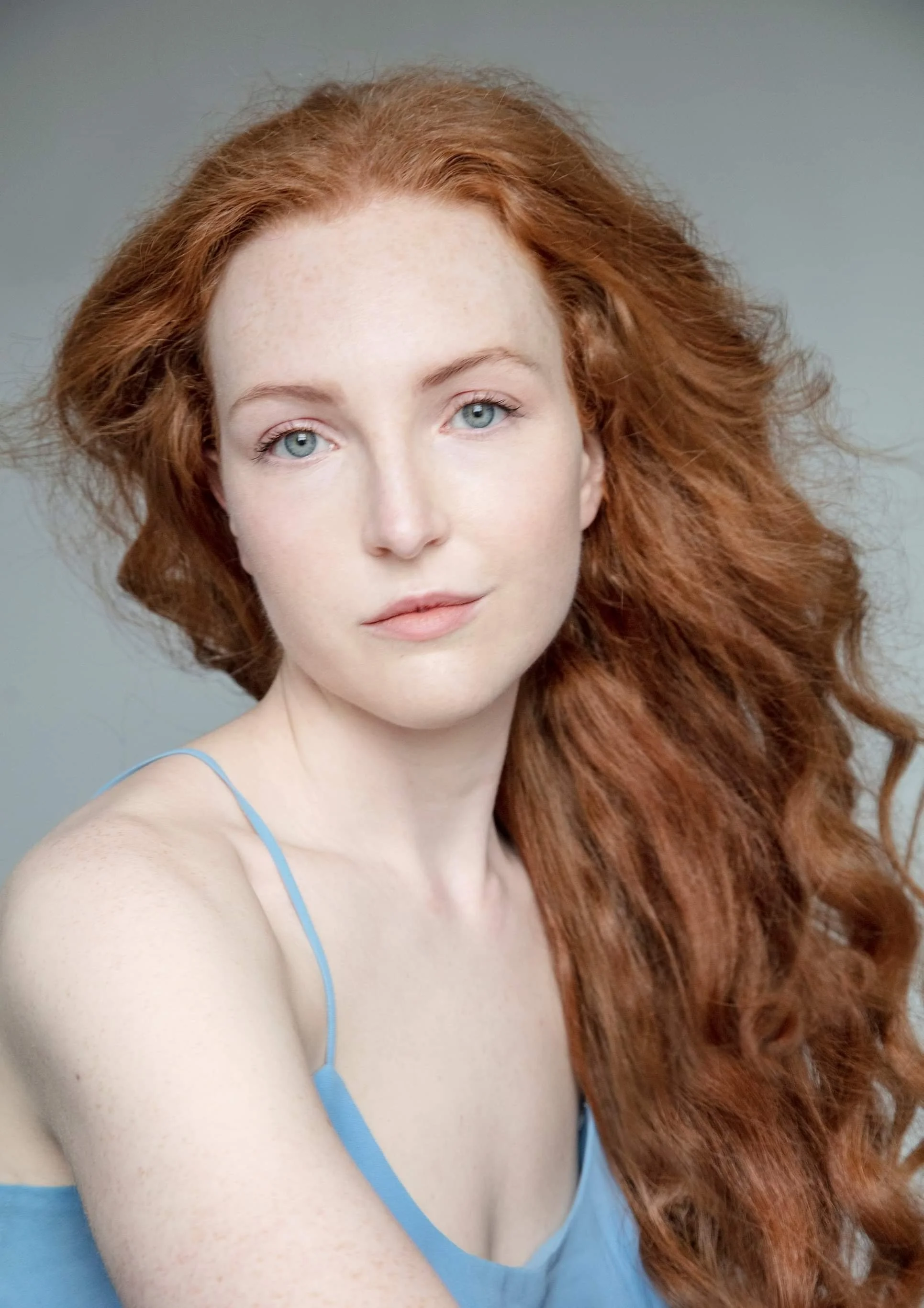A woman with long curly red hair looks at the camera - she has blue eyes and pale skin, and is wearing a light blue strap top. The background is grey