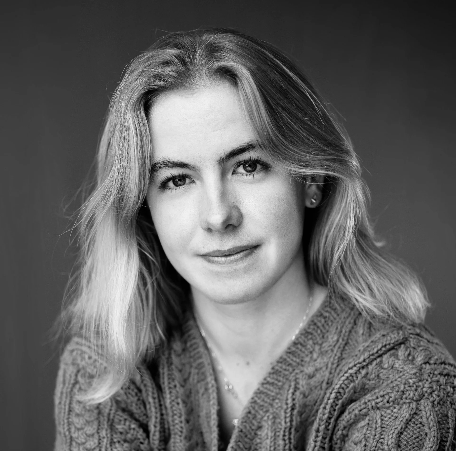 Black-and-white close-up of a young woman with shoulder-length blonde hair and light skin, wearing a knitted sweater, looking directly at the camera and smiling
