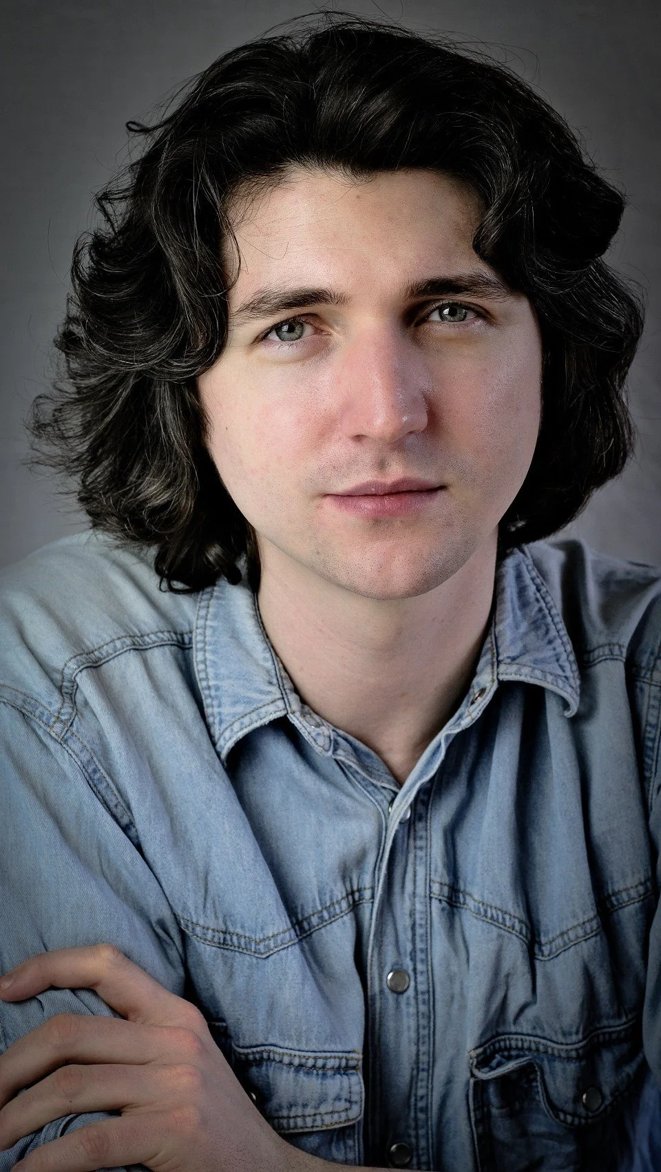 A young man looks directly at the camera - he has curly dark hair, pale skin and light eyes, and is wearing a light blue denim shirt.