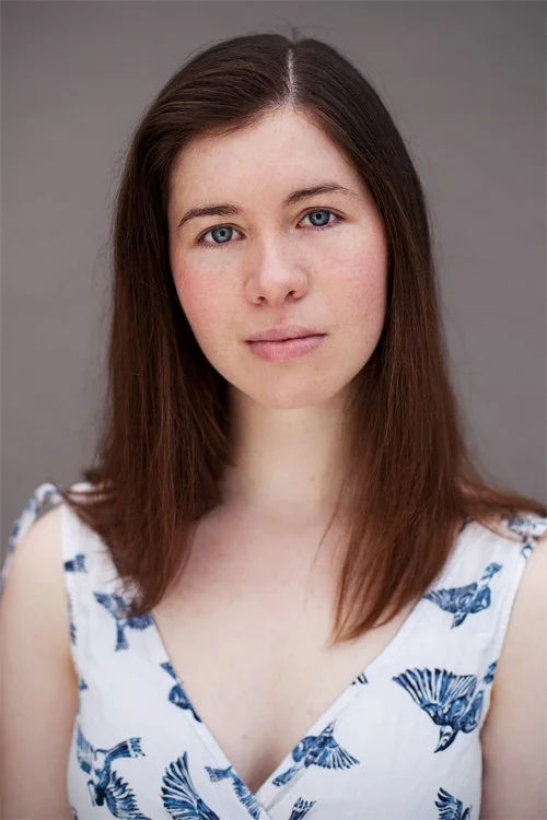 A young woman wearing a white and blue sleeveless dress looks directly at the camera. She has dark shoulder length hair, blue eyes, and pale skin. She is against a light grey backdrop