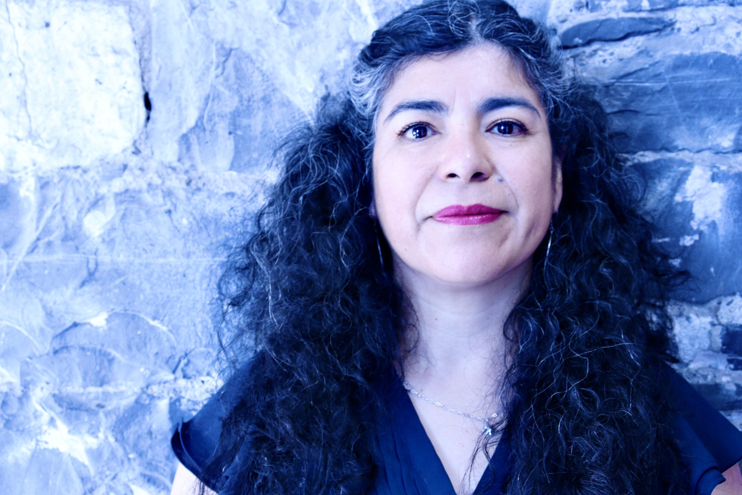 Close-up portrait of a woman with dark hair and dark eyes and red lipstick, wearing a dark v neck top and hoop earrings, looking at the camera, against a stone wall.