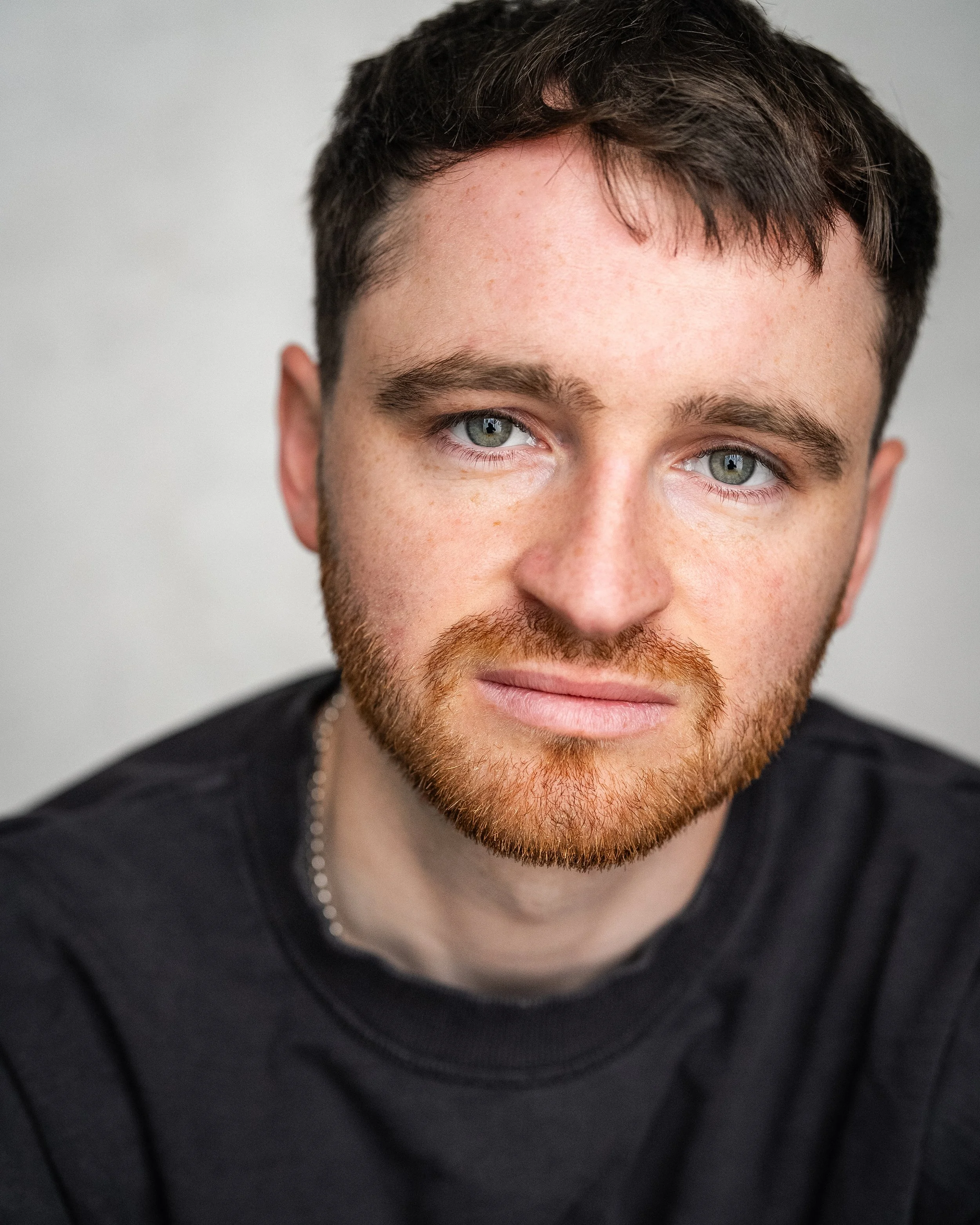A man with brown hair, hazel eyes, light skin and a reddish brown beard and moustache, wearing a black top, looks directly into the camera against a white wall