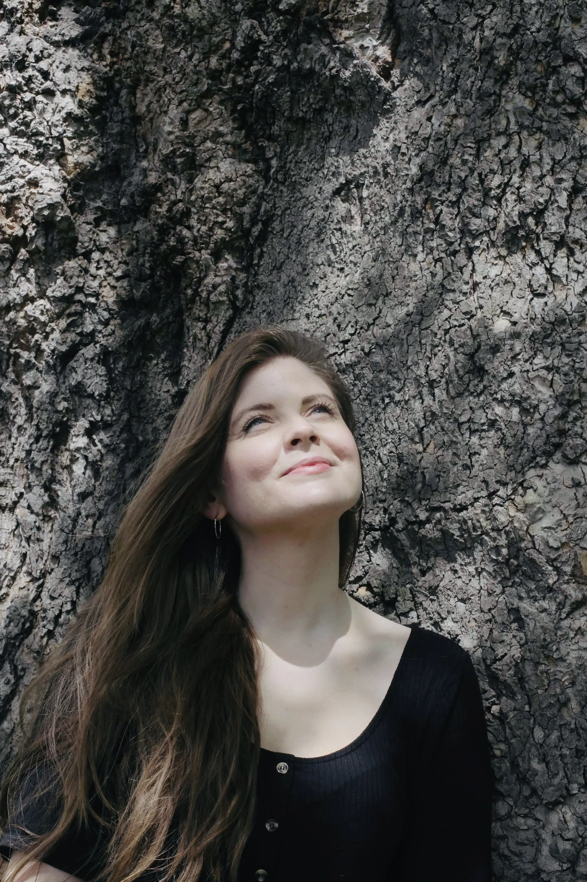 A young woman looks upwards. She has long brown hair and pale skin, and is wearing a black top. She's outside standing in dappled light against a textured stone background