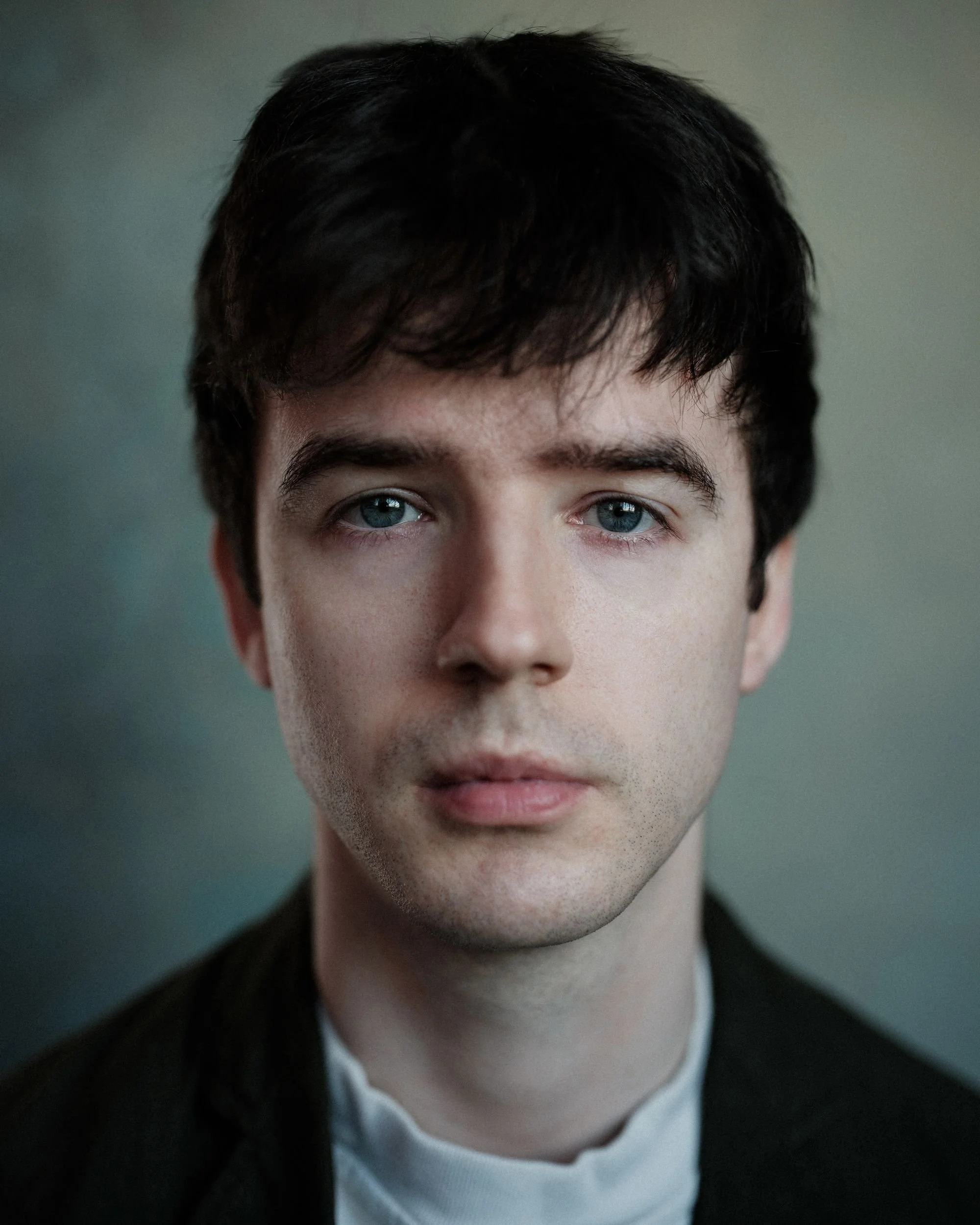 A young man with dark hair, blue eyes and pale skin looks directly at the camera. He is wearing a white tshirt and dark shirt, and is against a grey background