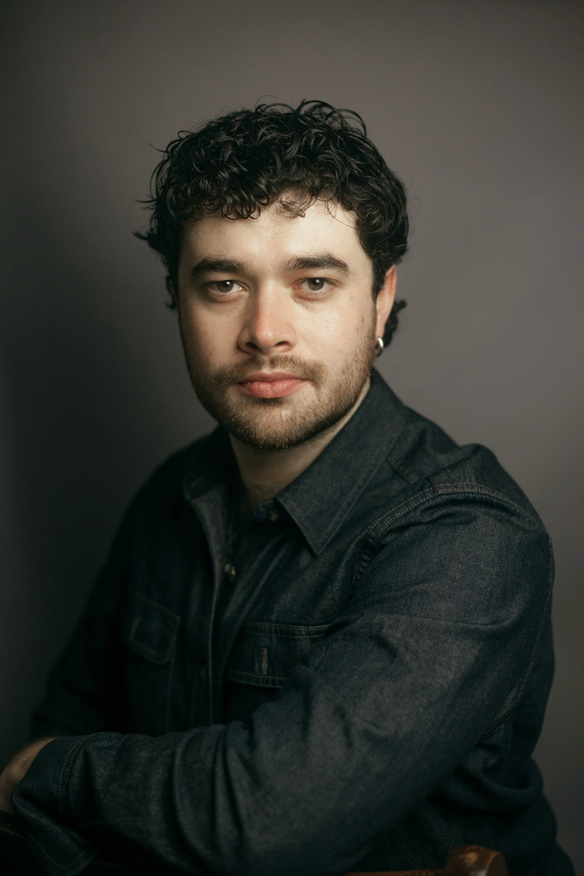 A young man with dark hair, dark eyes, and a beard and moustache, looks directly at the camera. He's wearing a black shirt and an earring, and is against a backdrop of dark grey