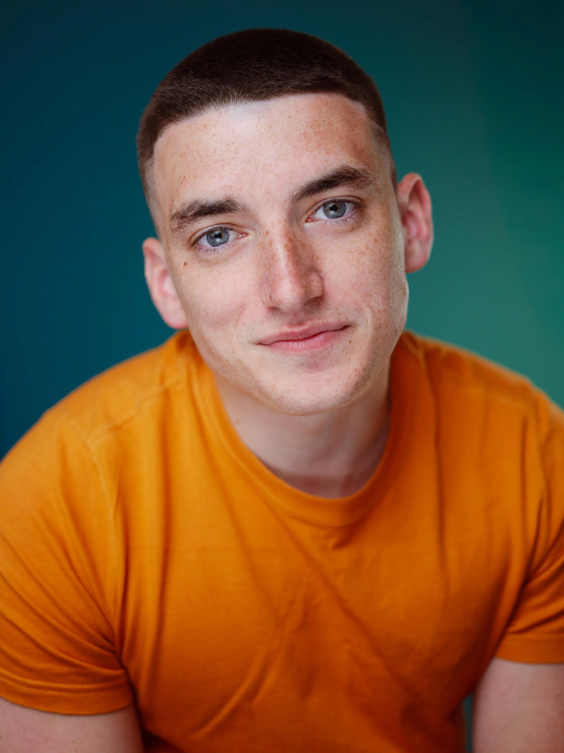 A young man with short dark brown hair smiles into the camera; he is wearing an orange tshirt and he's leaning forward slightly. The background is dark grey.