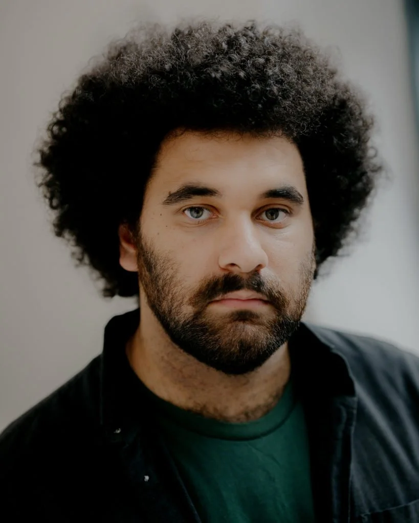A man with dark eyes, brown skin, and a beard/moustache and afro and wearing a dark green tshirt and black shirt looks directly at the camera against a white wall