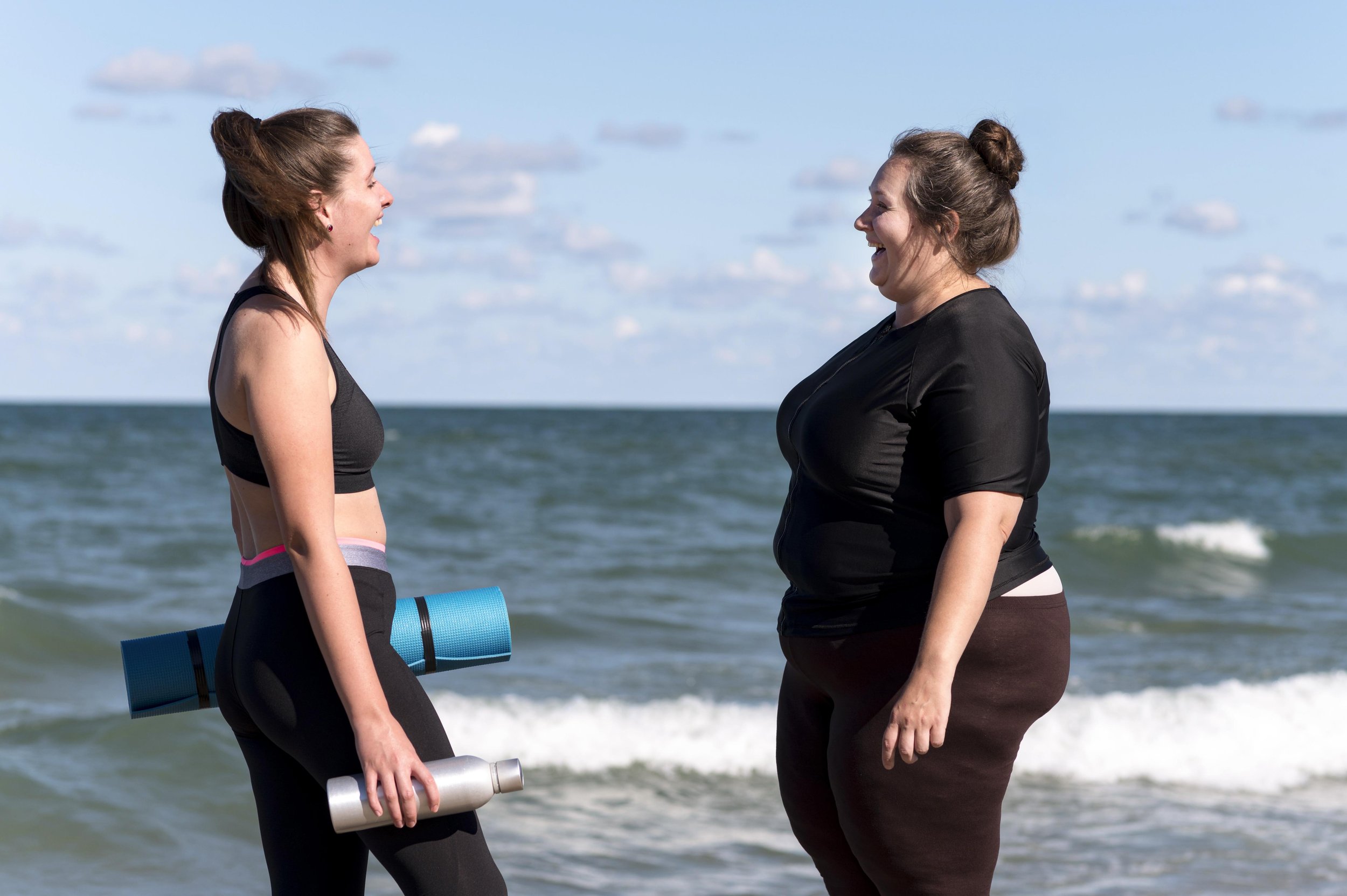 Two women laughing together at the beach with yoga mats and water bottles.