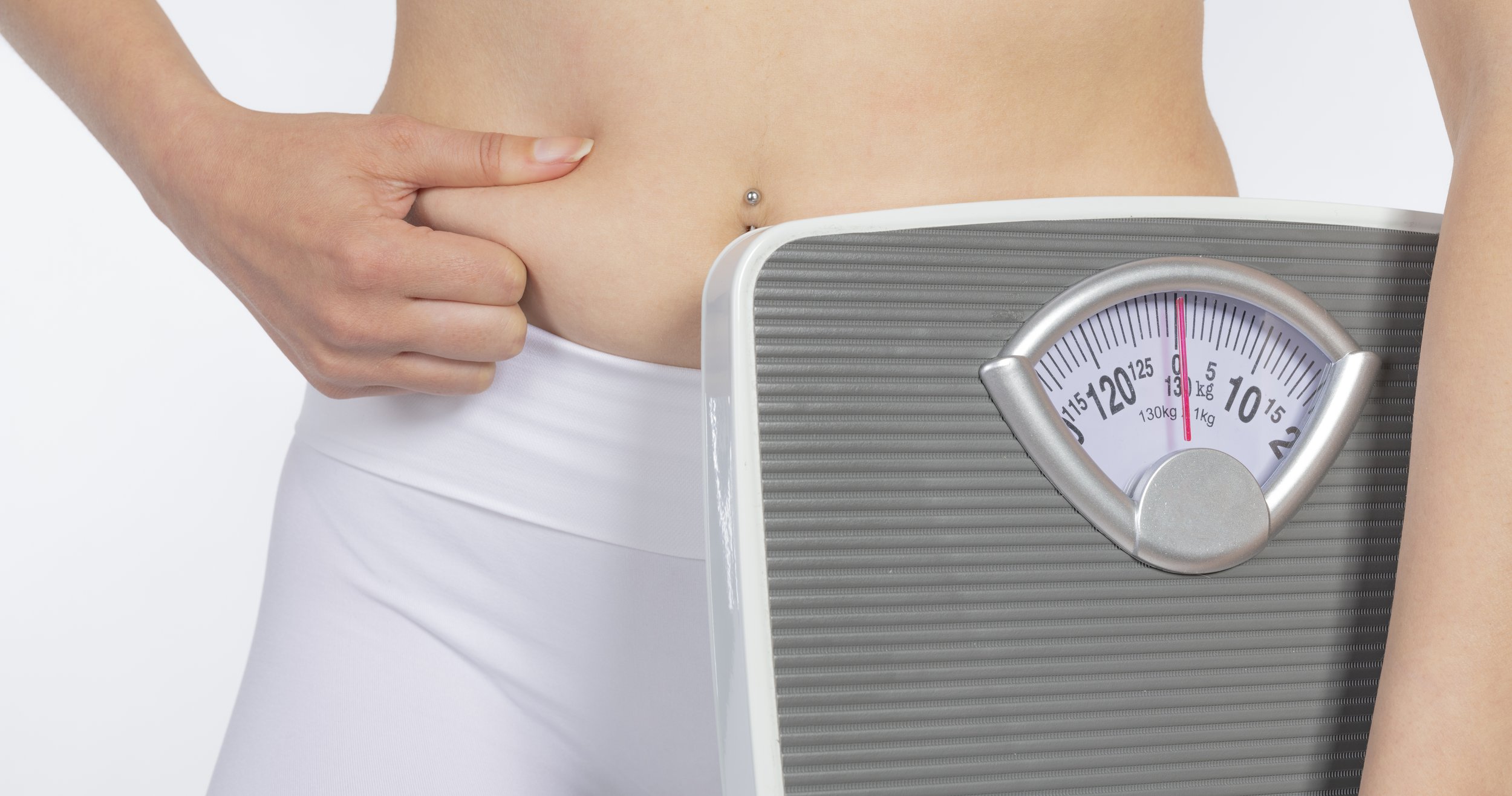 Woman pinching abdominal fat while standing next to a bathroom scale, illustrating weight loss and body composition on a white background.