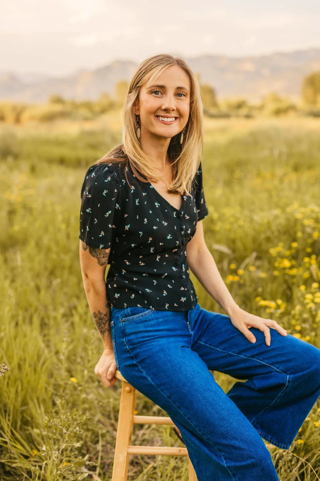 A woman with blonde hair and tattoos, wearing a black floral shirt and blue jeans, sitting on a wooden stool in a field of yellow flowers with mountains in the background.