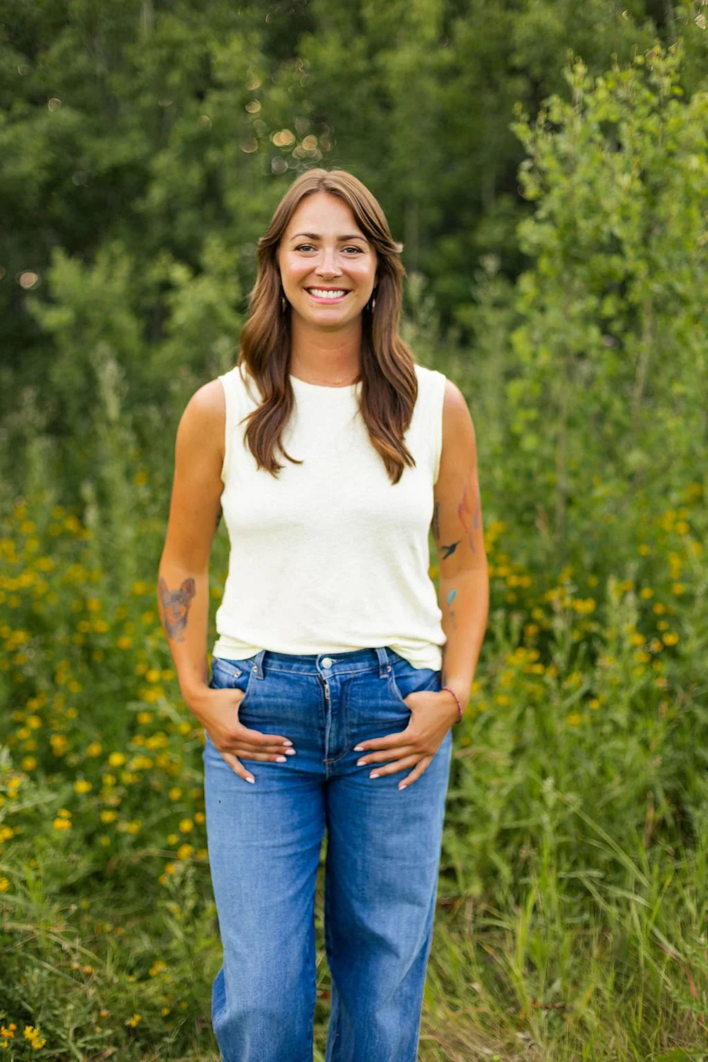 A woman standing outdoors in a green, natural setting, smiling at the camera, wearing a sleeveless white top and blue jeans.