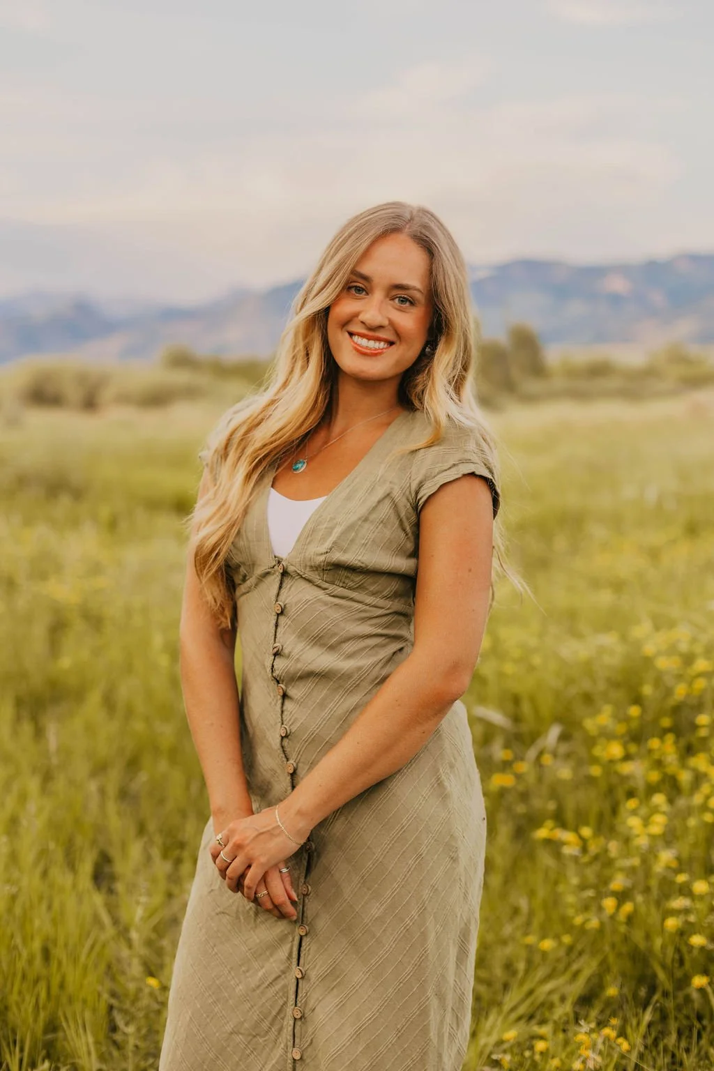 A smiling woman with long blonde hair standing in a field of yellow flowers with mountains in the background.