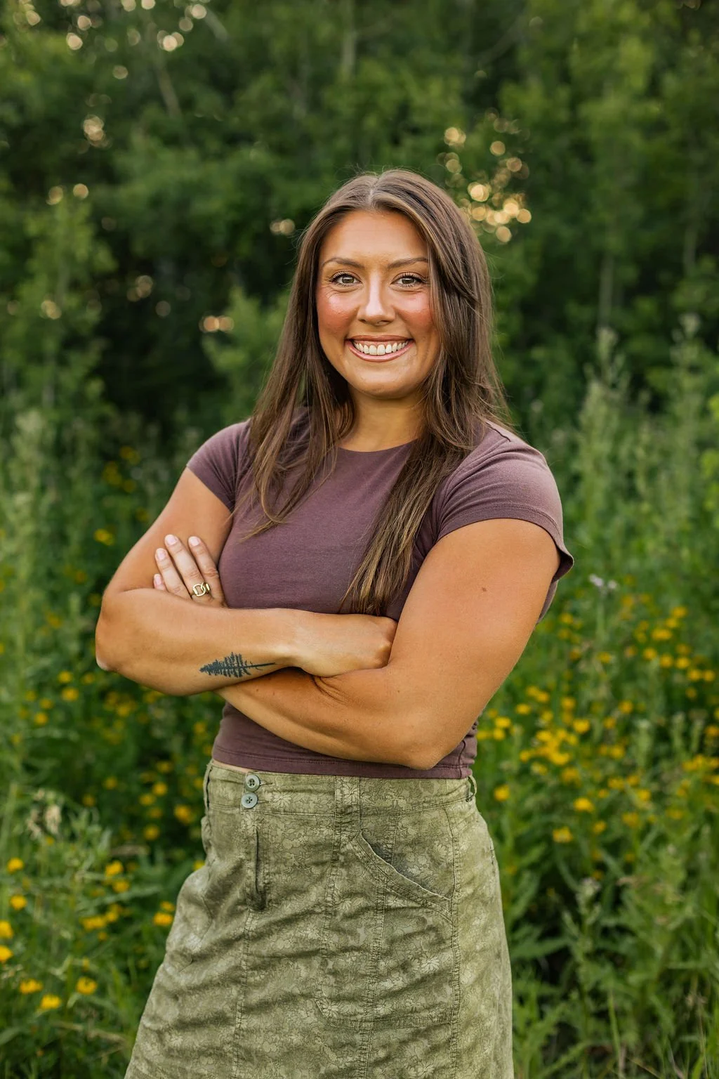 A woman with long brown hair standing outdoors in a green field, smiling with arms crossed, wearing a brown t-shirt and a patterned green skirt, with a tattoo of trees on her left forearm.