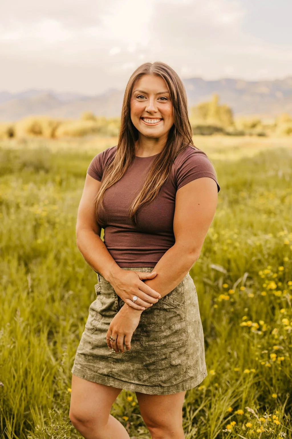 A woman with long brown hair smiling in a field of yellow flowers with mountains in the background.