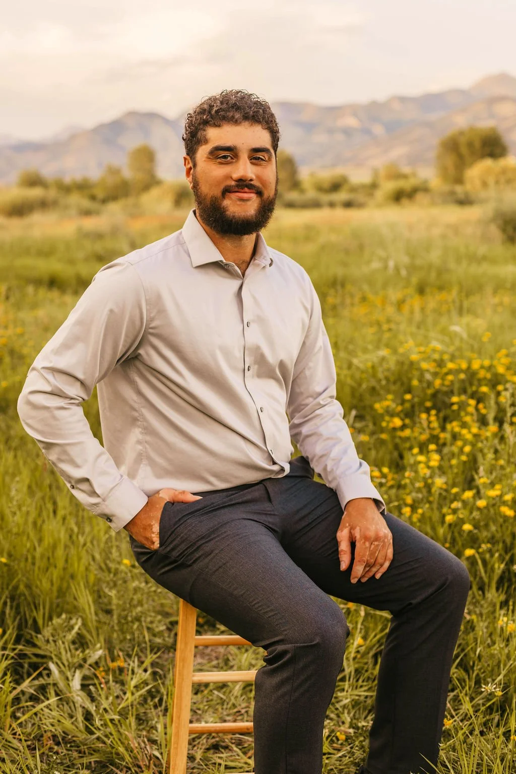 A man with curly hair and a beard, wearing a light gray dress shirt and dark pants, sitting on a wooden stool in a field of yellow wildflowers with mountains in the background during sunset.