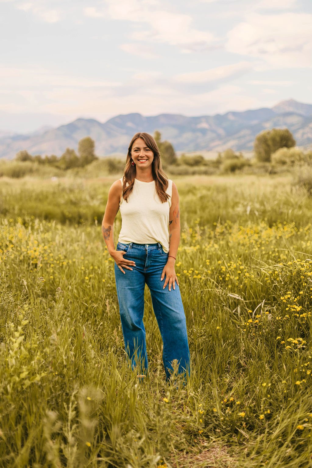 A woman standing in a field of yellow wildflowers with mountains in the background during daytime.