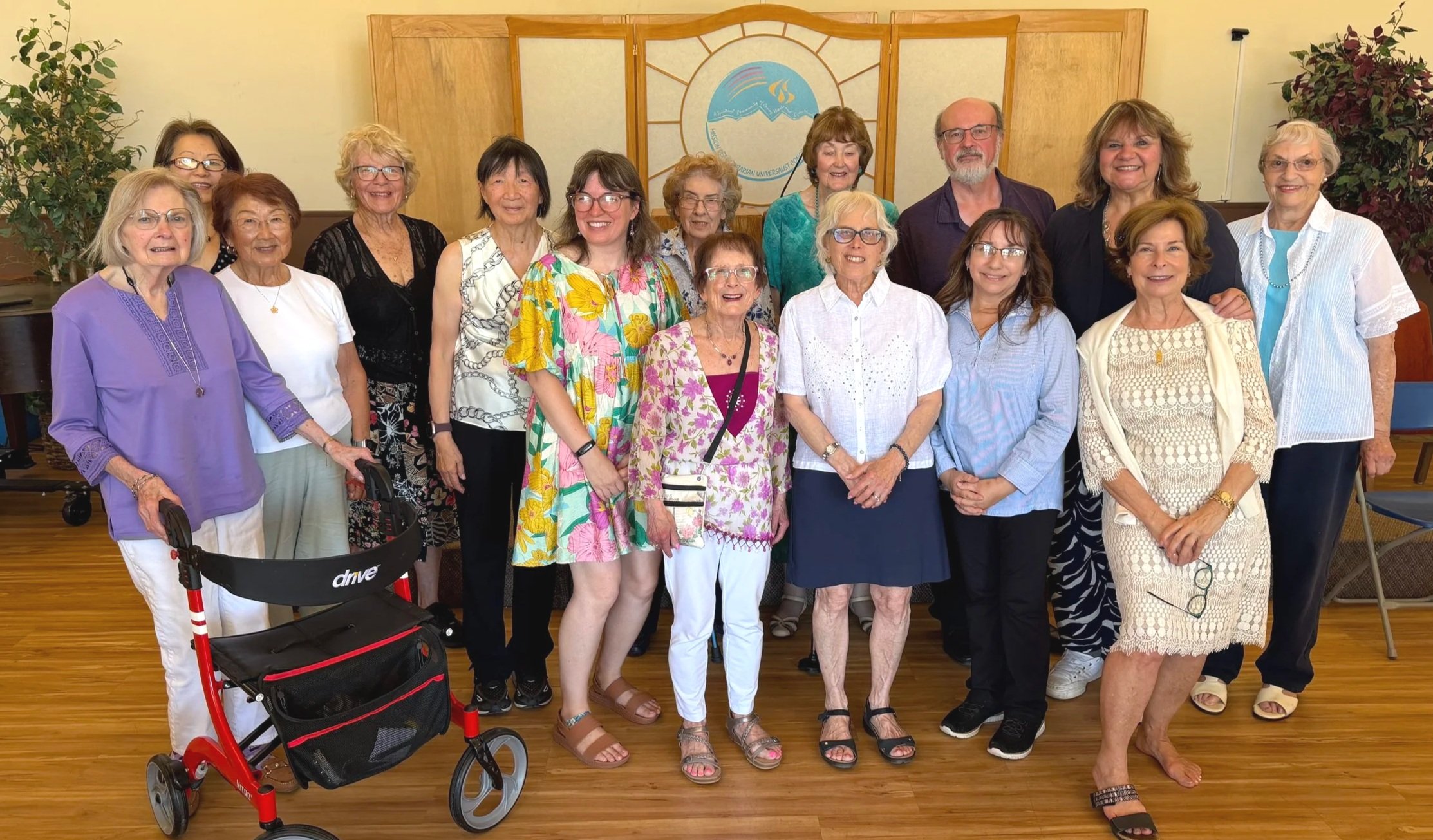 A group of sixteen women and one man posing together indoors in front of a wooden wall with a sign and a picture of mountains and a rainbow. The women are smiling and wearing casual summer clothes, with some wearing glasses. One woman has a walker, and there is a red and black stroller in the foreground.